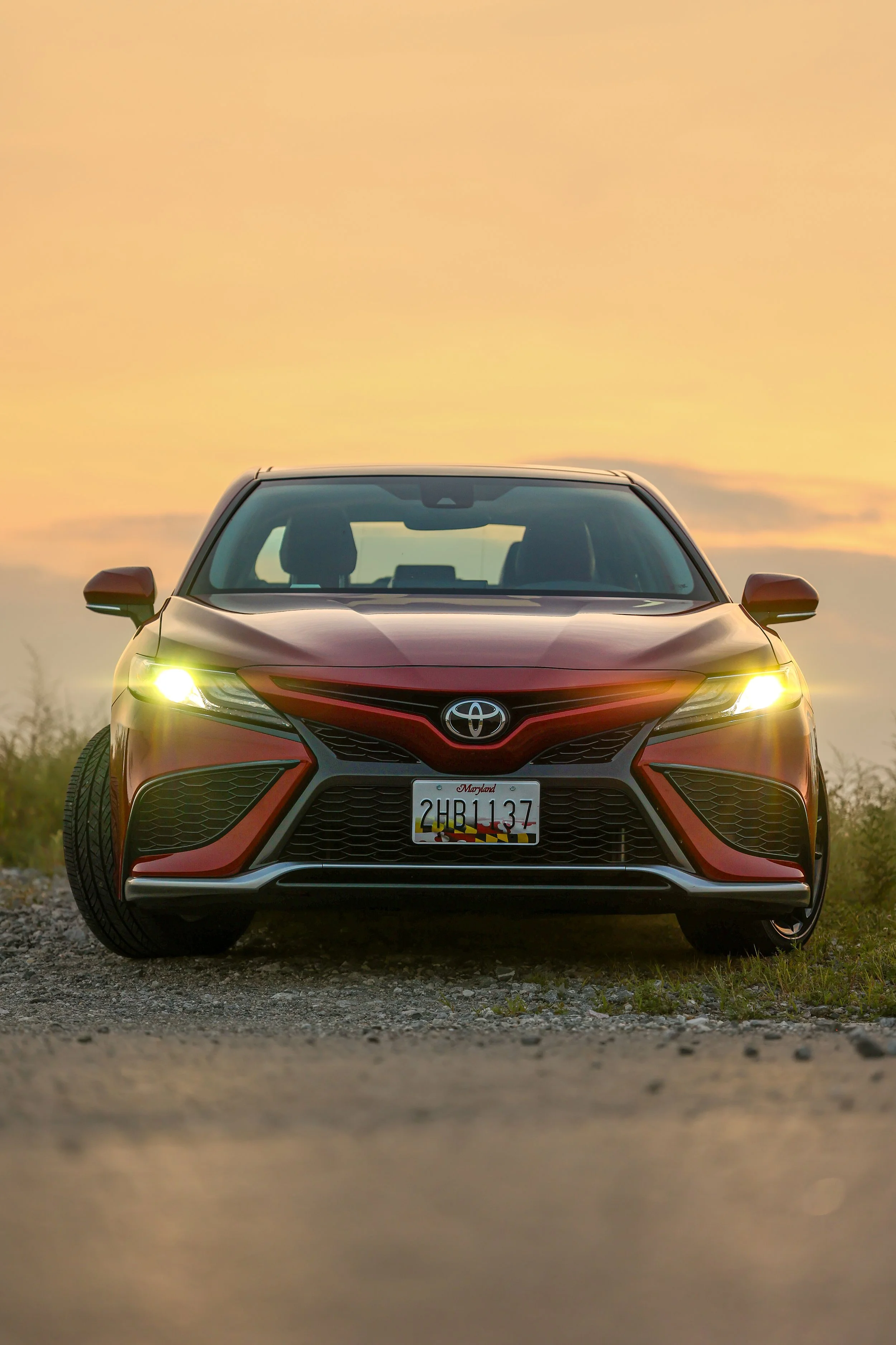 Front view of a red Toyota sedan car with headlights on, parked on a gravel surface during sunset, with a blurred background of a yellow-orange sky.