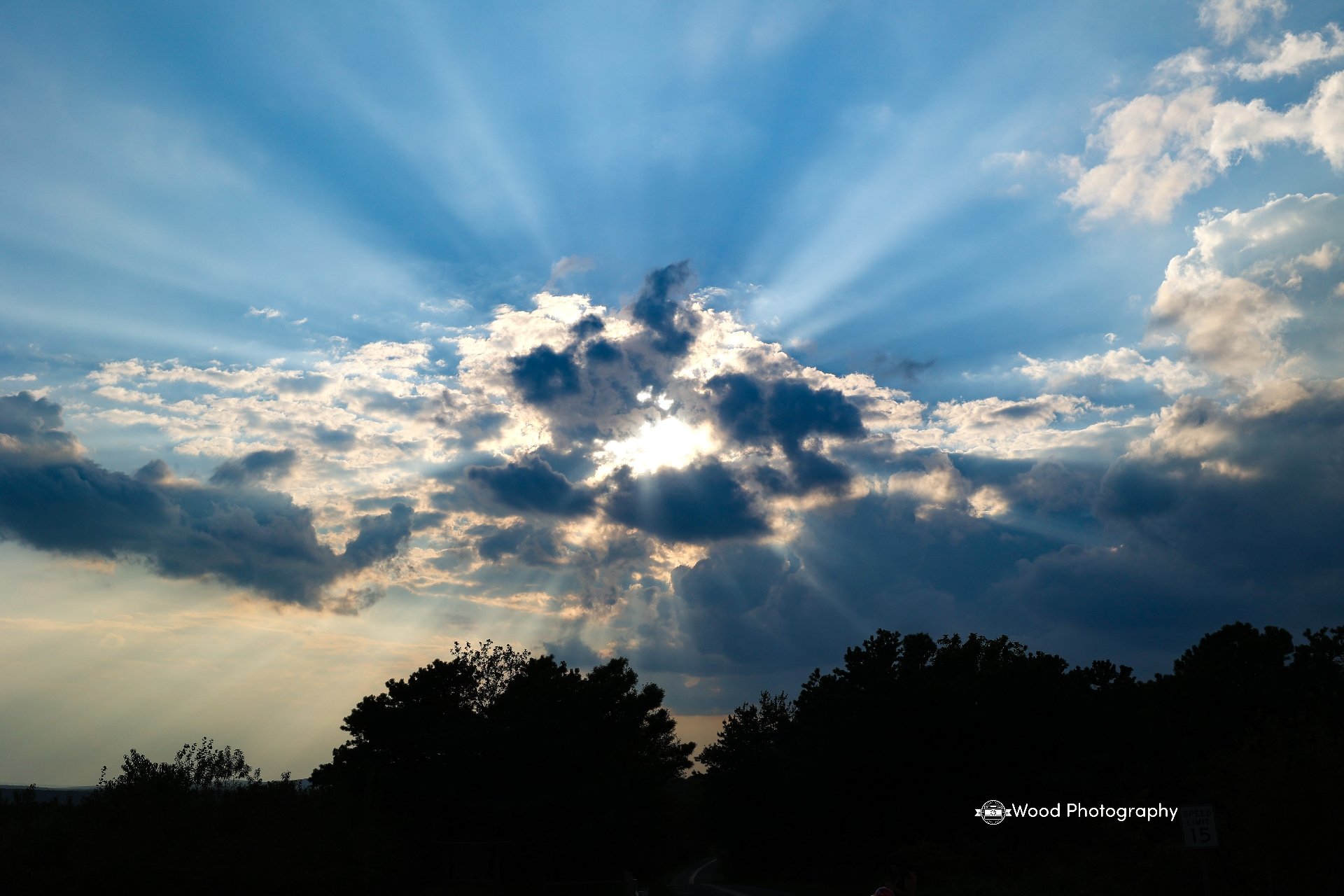 Sky with clouds and sunlight streaming through, silhouetted treetops at the horizon.