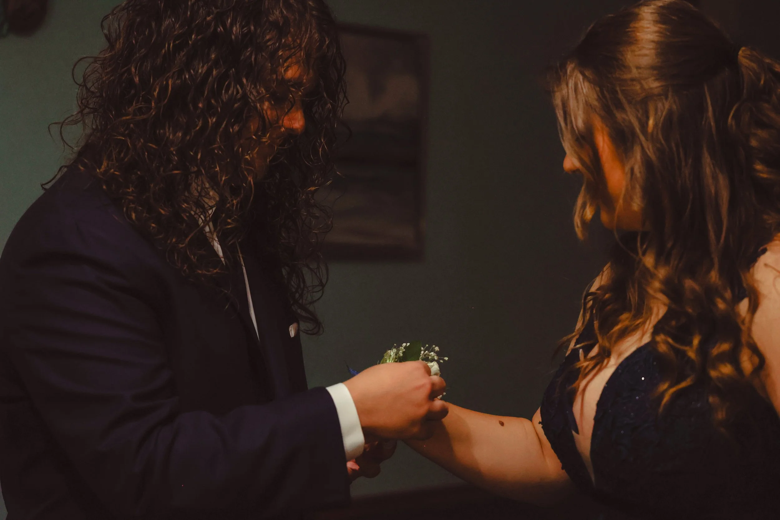 A woman in a dark, sleeveless dress is receiving a wedding ring during a ceremony, with a man in a black suit and white shirt presenting the ring. They are holding hands, and a small bouquet is visible.