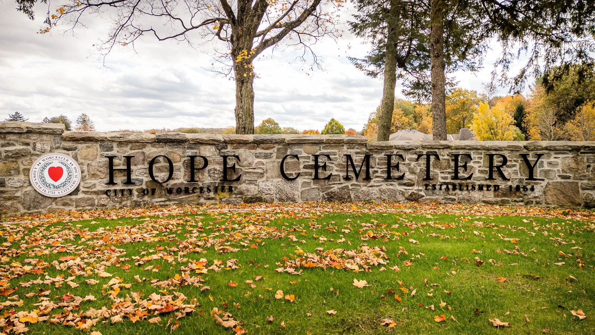 Stone sign reading 'Hope Cemetery, City of Worcester, Established 1854' with a Worcester city seal featuring a red heart, surrounded by fall foliage and trees with autumn leaves.
