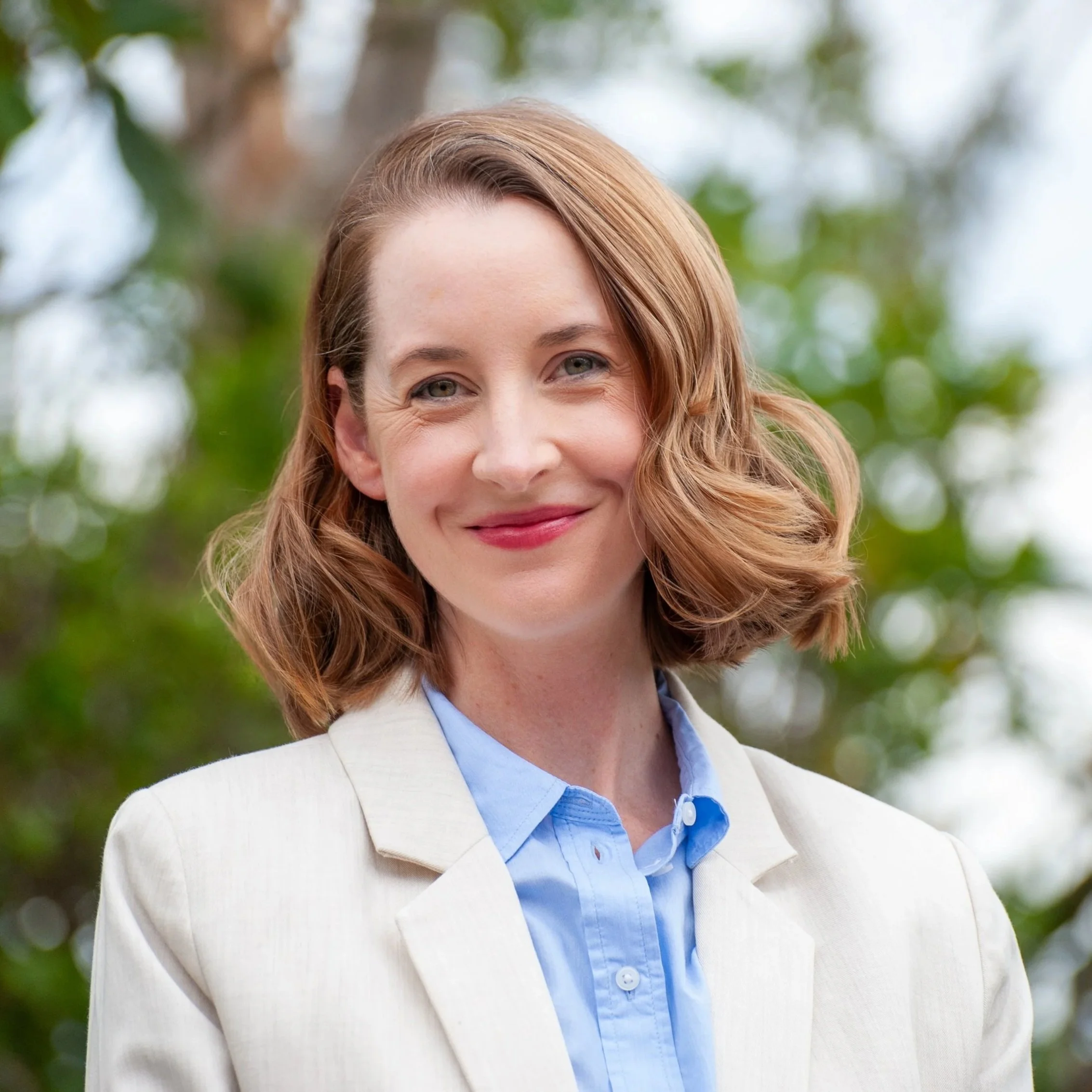 A woman with shoulder-length red hair, smiling, wearing a light-colored blazer over a blue collared shirt, outdoors with green trees in the background.