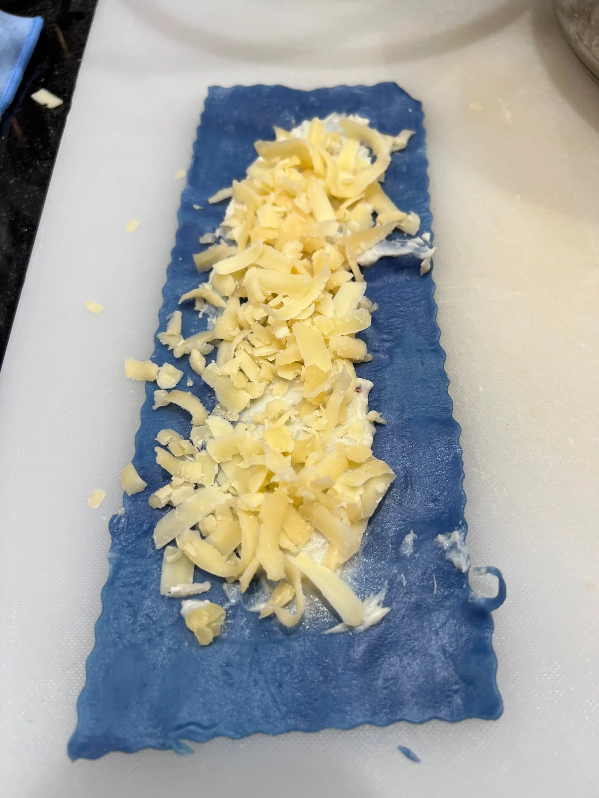 Unrolled blue pasta sheet topped with shredded cheese on a white cutting board.