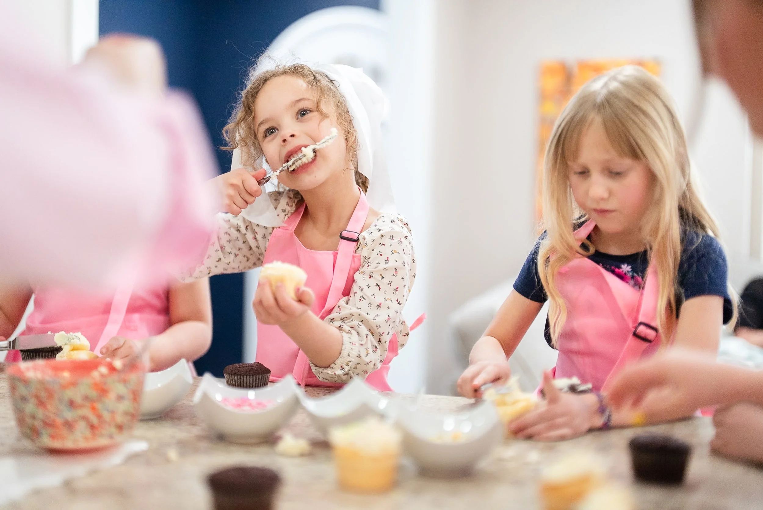 Two young girls wearing pink aprons baking cupcakes in a kitchen.