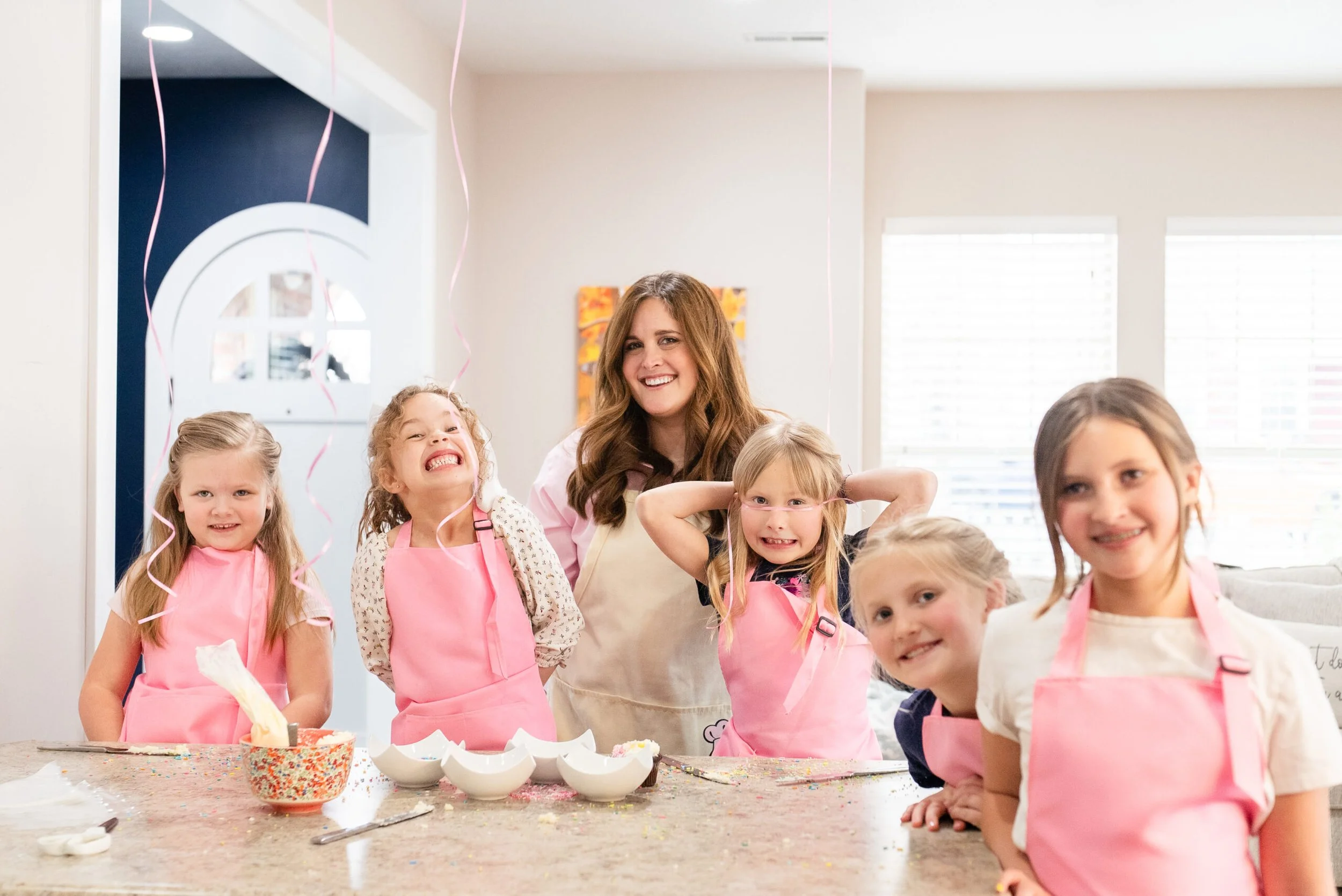 A group of young girls and a woman, likely a teacher or parent, celebrating a birthday in a kitchen, with pink aprons, balloons, and a decorated table.