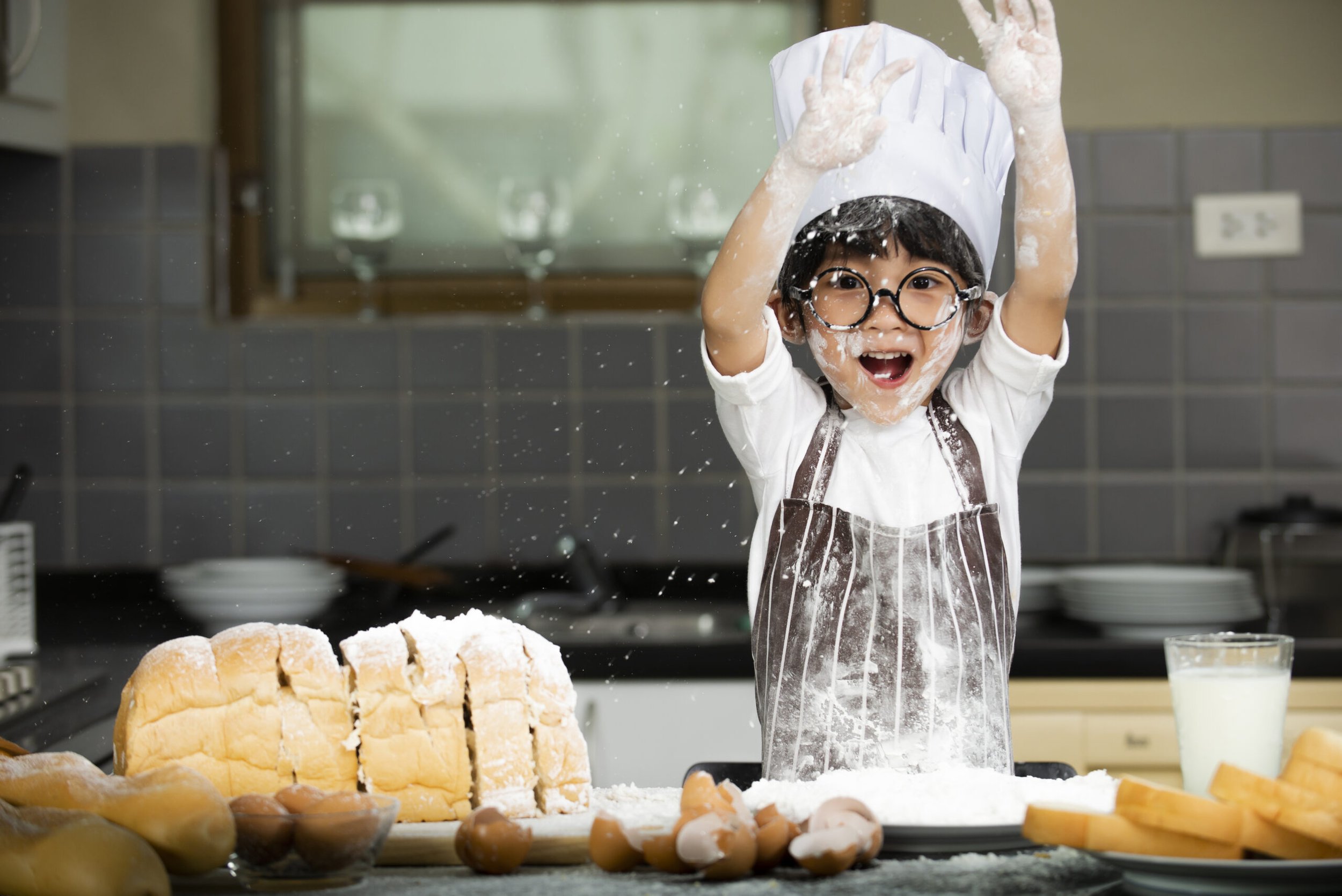 A young boy wearing glasses, a chef's hat, and an apron is happily throwing flour in the air in a kitchen. There is bread, eggs, and a glass of milk on the counter, indicating baking activities.