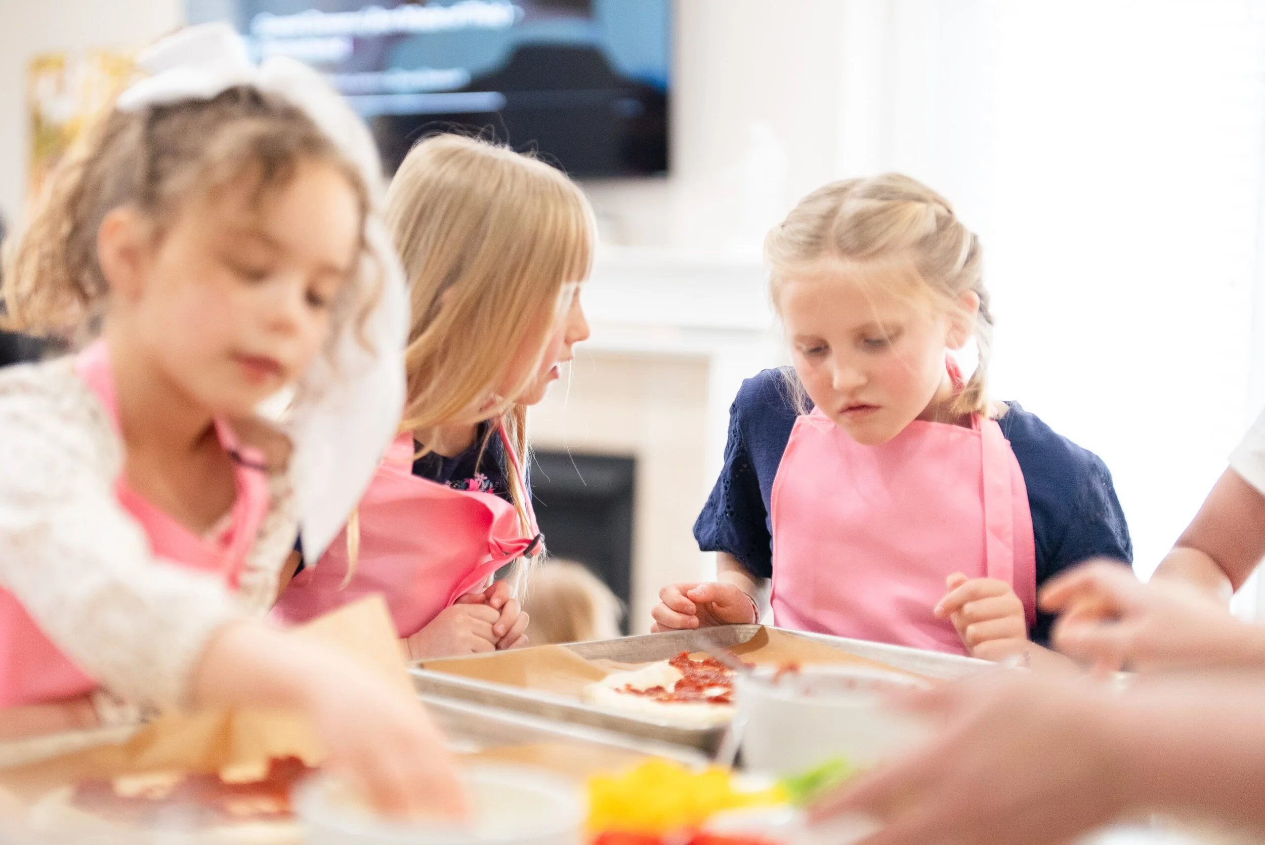 Three young girls wearing pink aprons and concentrating on making pizza in a kitchen.