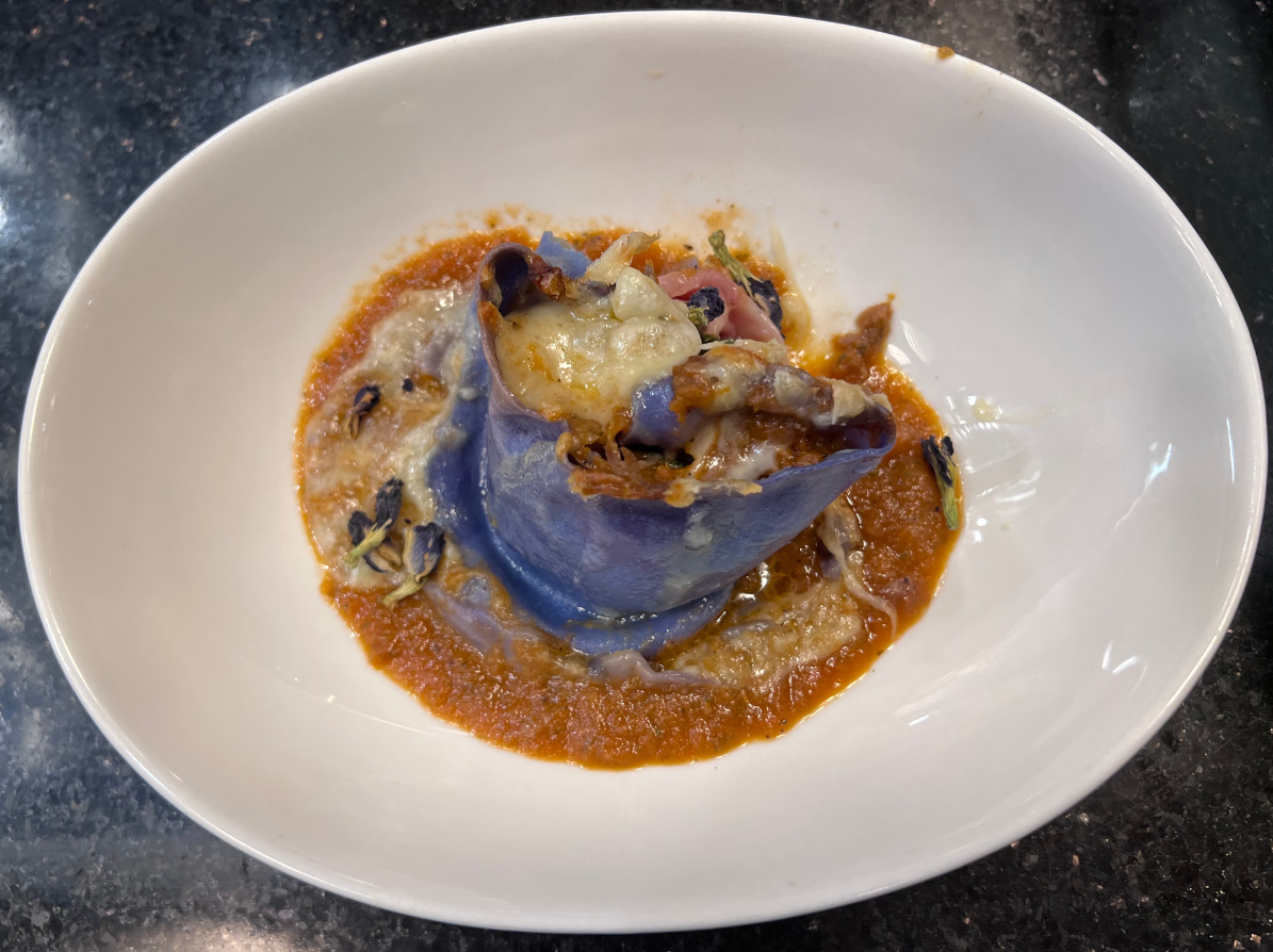 Blue pasta roll topped with cheese and herbs in tomato sauce on a white oval plate.