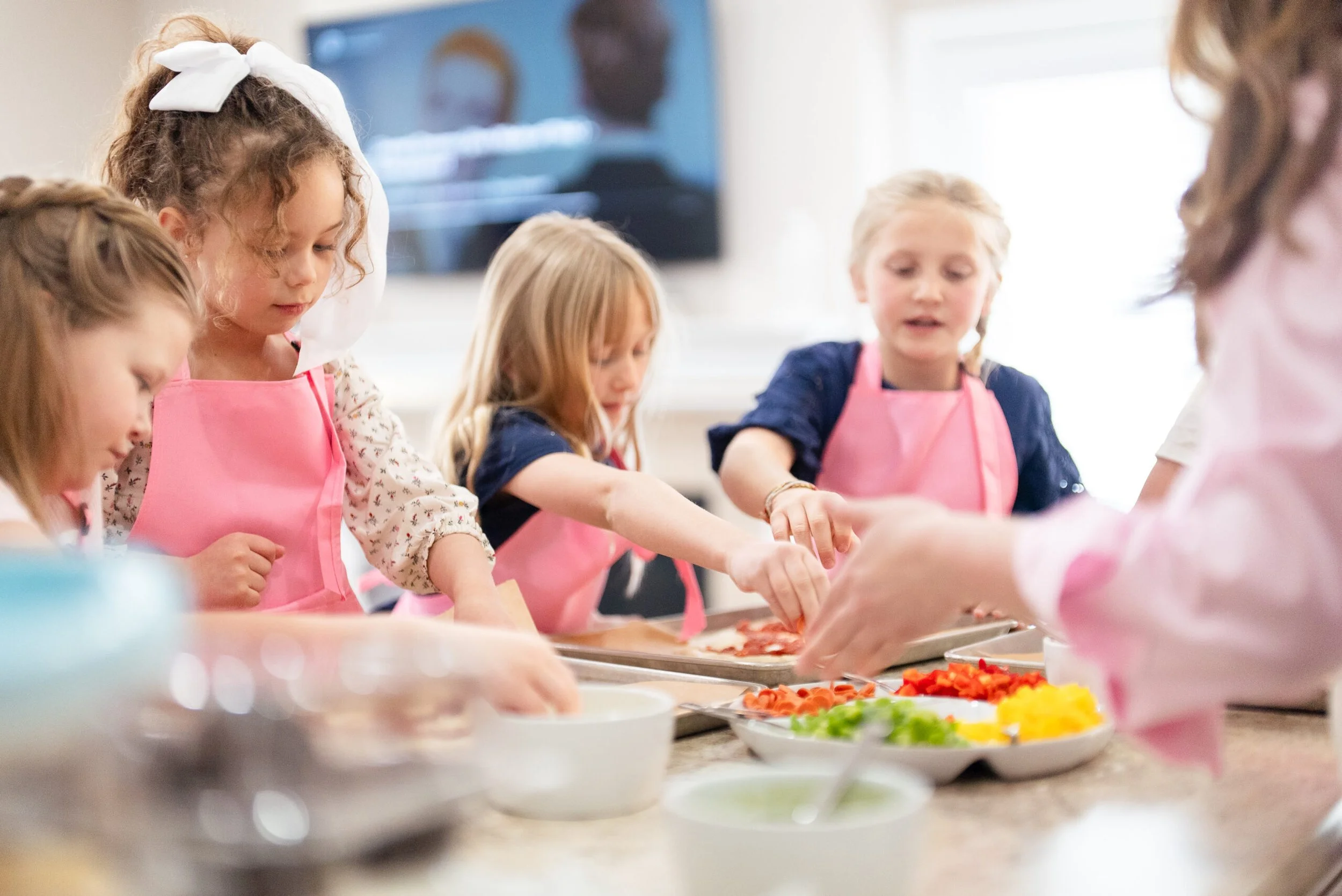 Children wearing pink aprons preparing food together in a kitchen.