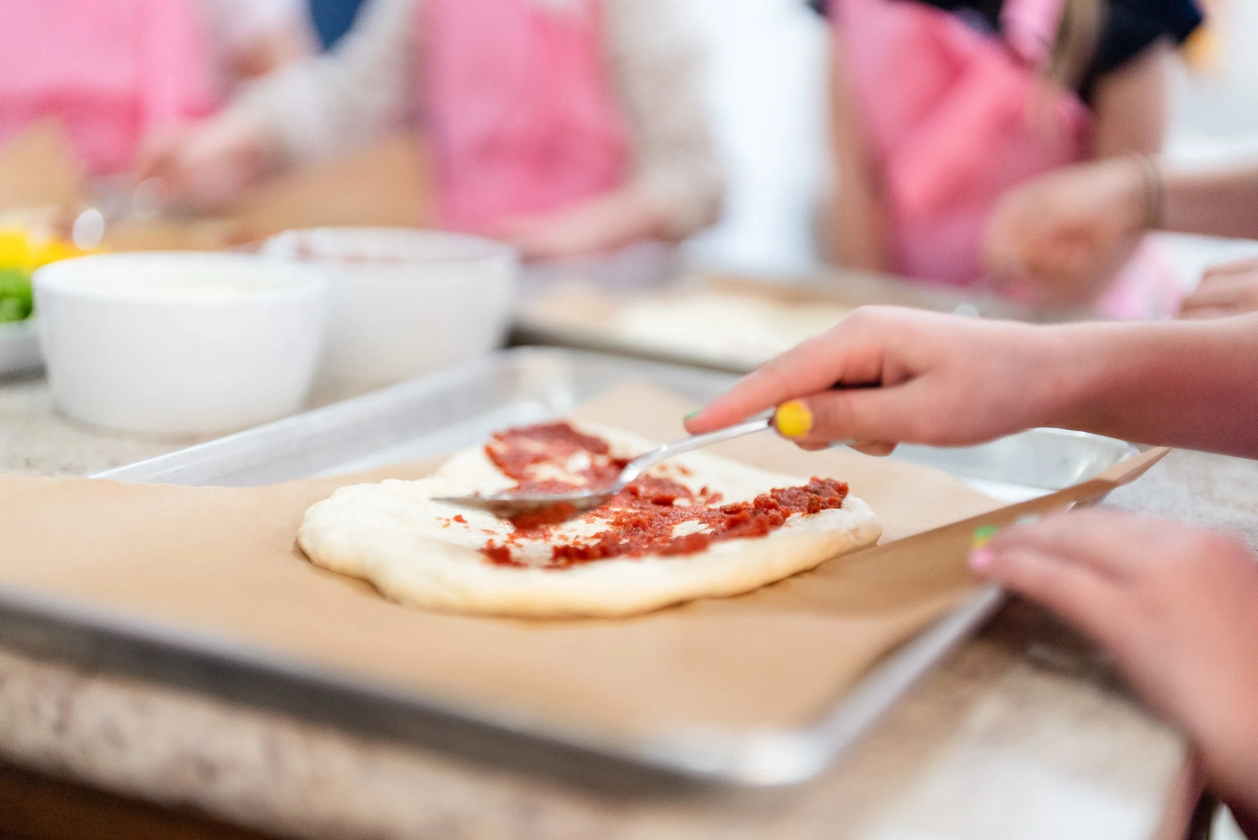 Person spreading tomato sauce on pizza dough with a spoon in a kitchen setting with other children in pink aprons in the background.