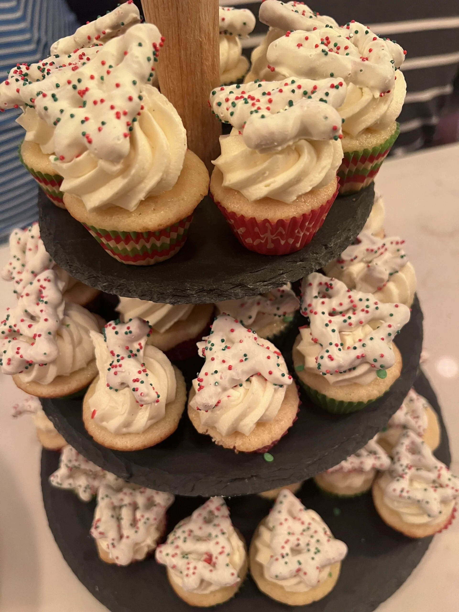 Assorted Christmas-themed cupcakes with white frosting and red and green sprinkles on a black tiered stand.