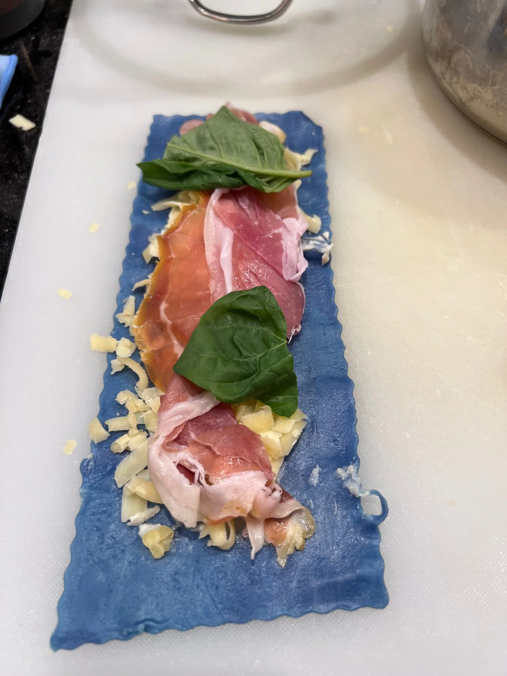 Blue pasta sheet topped with cheese, slices of prosciutto, and fresh basil leaves.