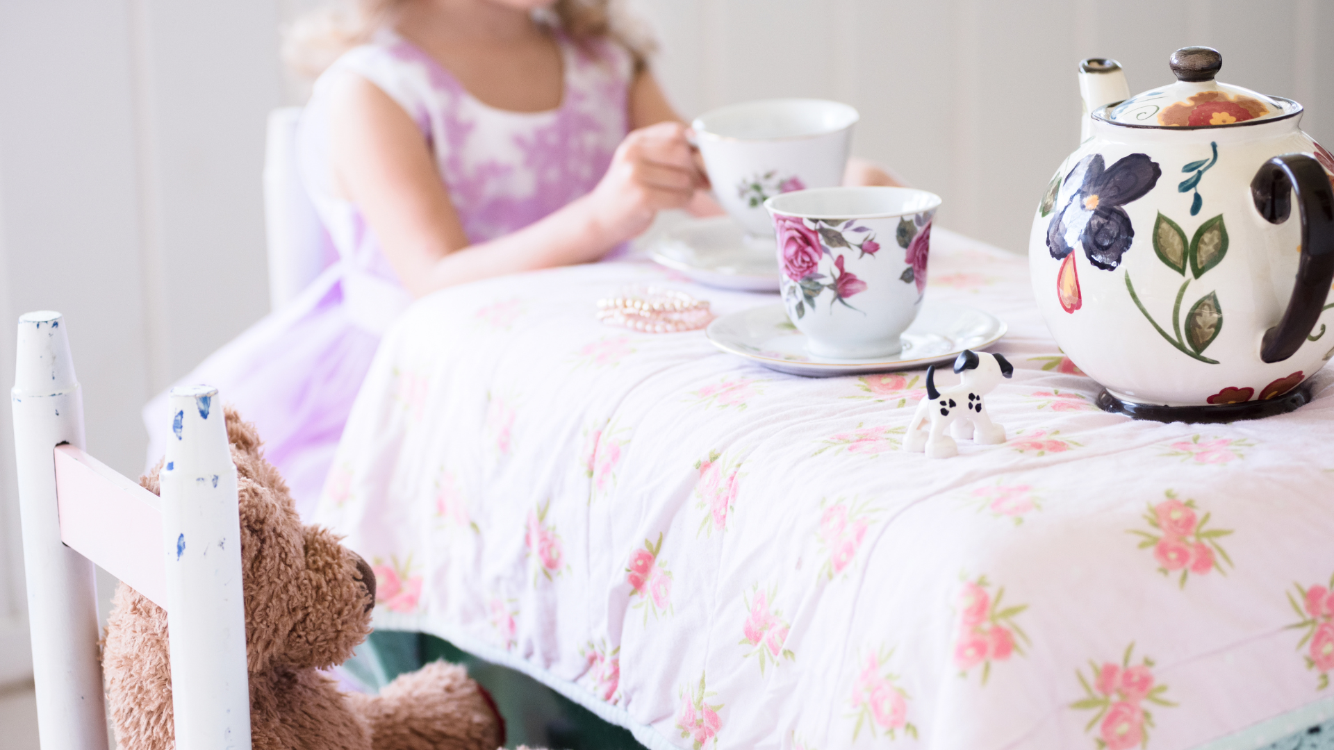 A young girl sitting at a bed with a floral tablecloth, holding a tea cup, with a teapot, a bowl, a small dog figurine, a pearl necklace, and a teddy bear nearby.