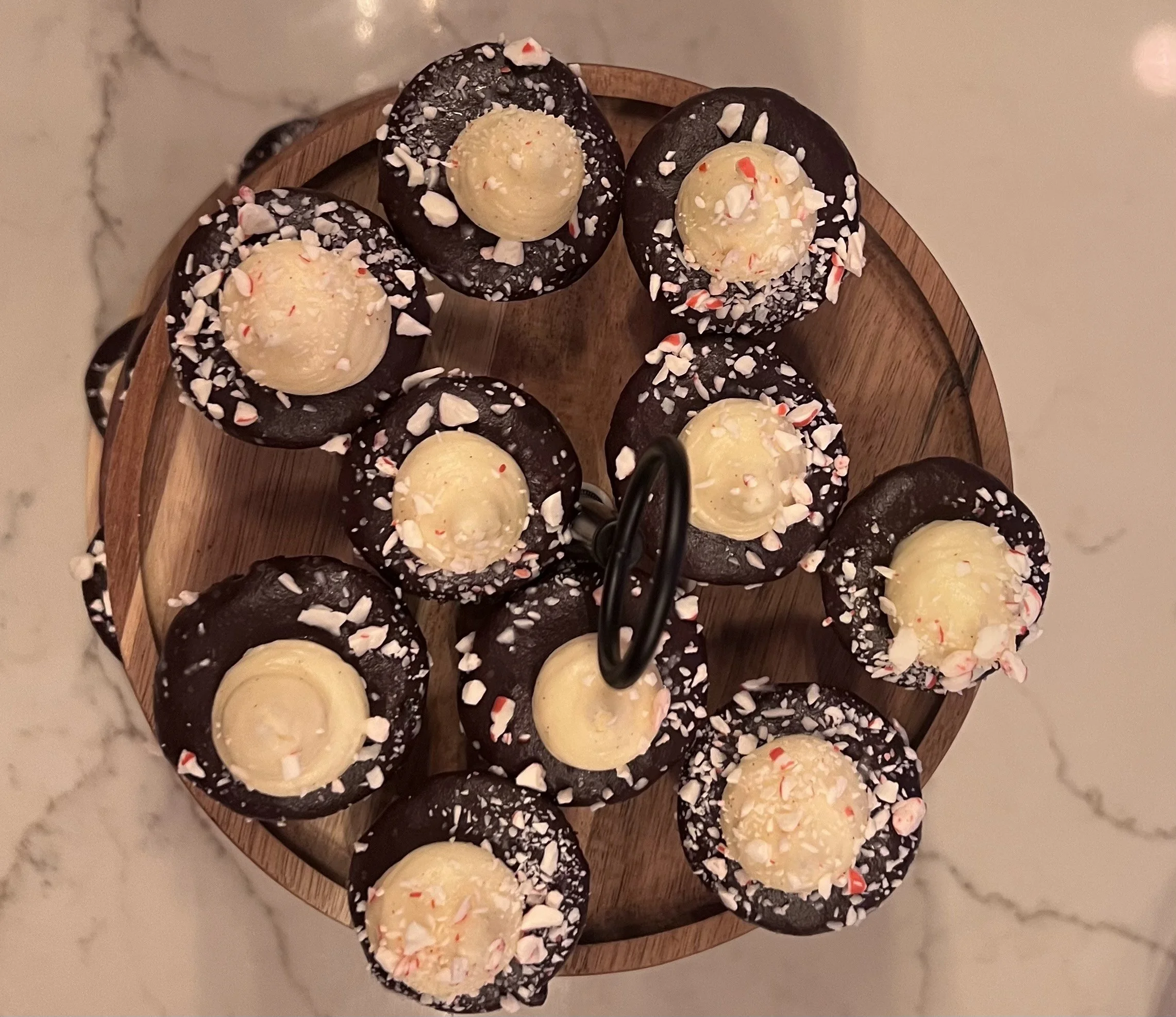 Chocolate cookies topped with white chocolate and crushed peppermint candies on a round wooden serving tray.