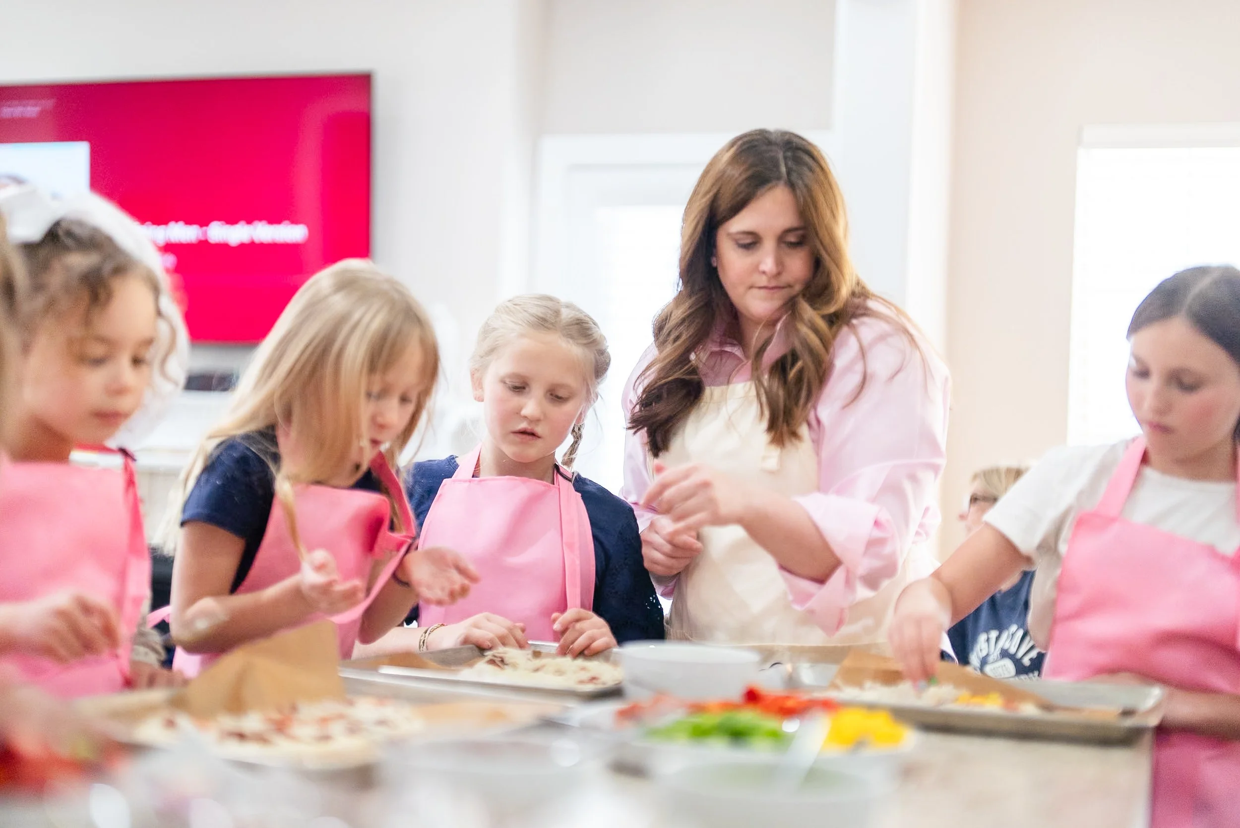 A woman teaching children how to make pizzas in a kitchen classroom, with kids wearing pink aprons around a table with pizza ingredients.