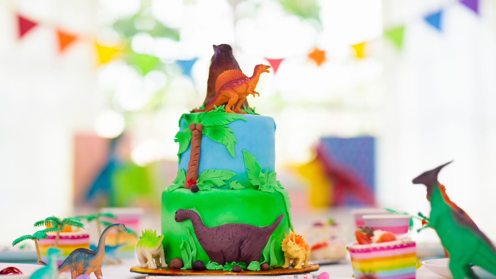 Colorful two-tiered dinosaur-themed birthday cake with green, blue, and purple fondant, decorated with dinosaur figurines, tropical leaves, and bunting, on a table with cupcakes and decorations.
