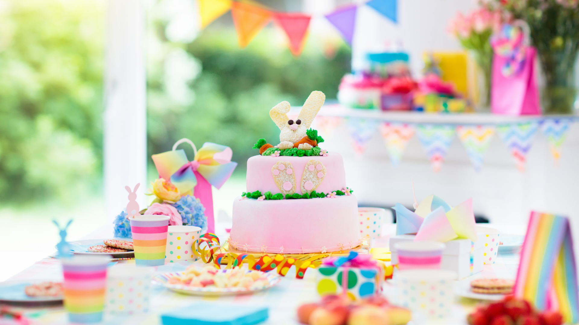 Decorated pink birthday cake with bunny topper, surrounded by party cups, plates, wrapping paper, and balloons, with colorful bunting hanging in the background at a festive celebration.