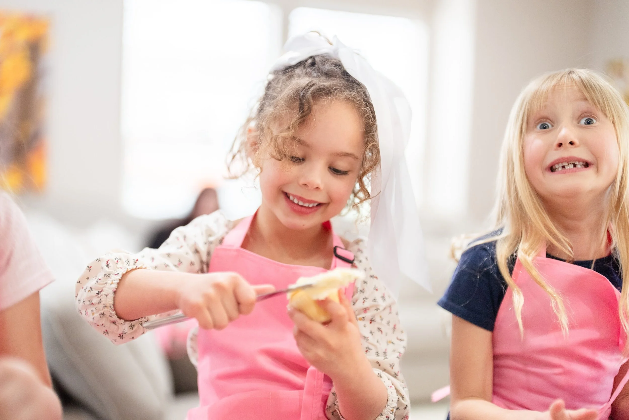 Two young girls wearing pink aprons in a kitchen, one with curly hair and a large white bow, spreading frosting on a cupcake with a knife, smiling and having fun.