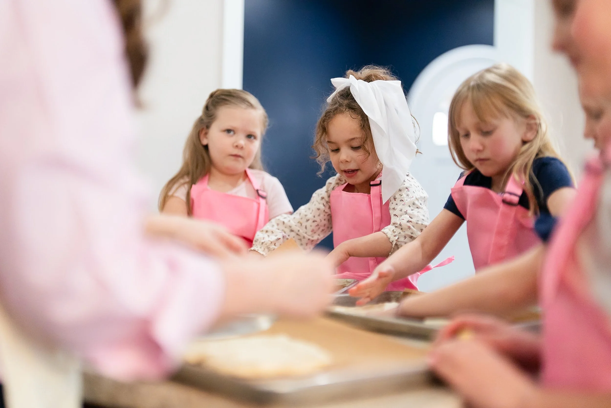 Group of young children baking together, wearing pink aprons in a kitchen.