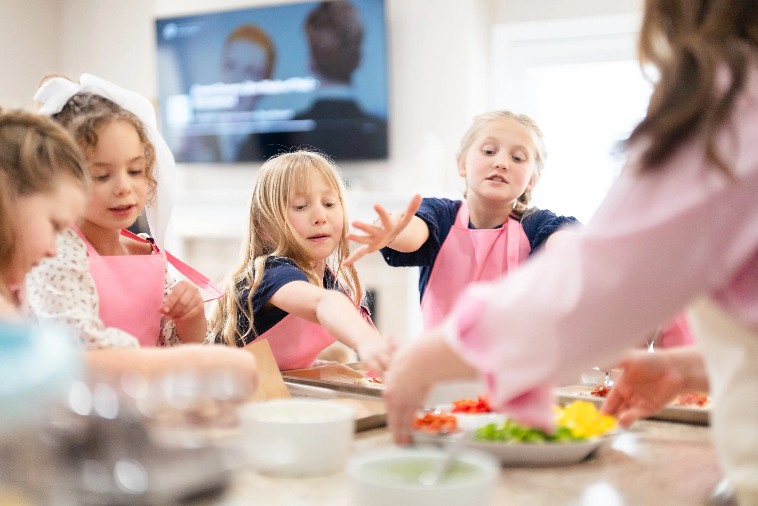 Children wearing pink aprons participating in a cooking or baking class, reaching for ingredients on a kitchen counter.