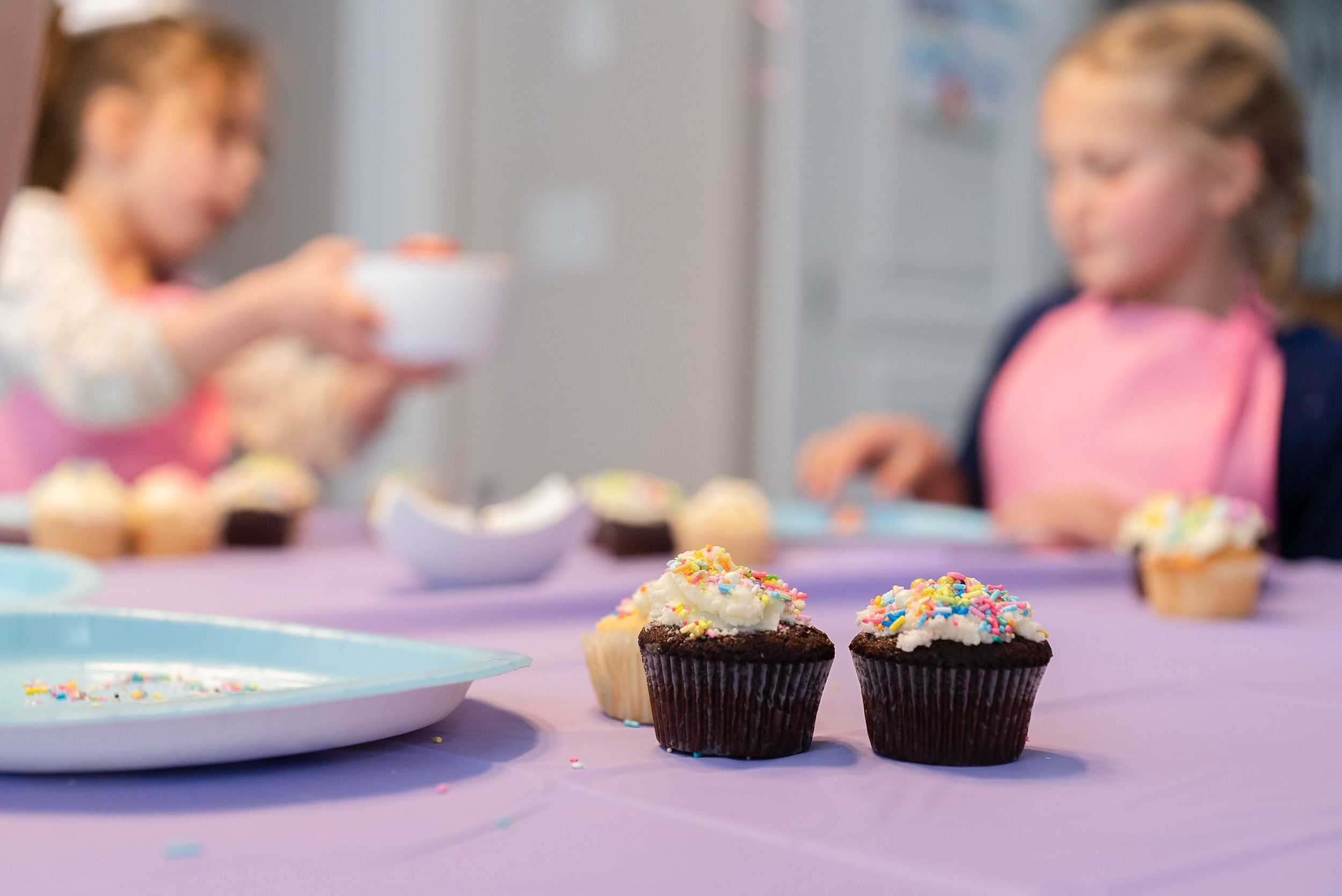 Two cupcakes with white frosting and colorful sprinkles on a purple table after a birthday party.