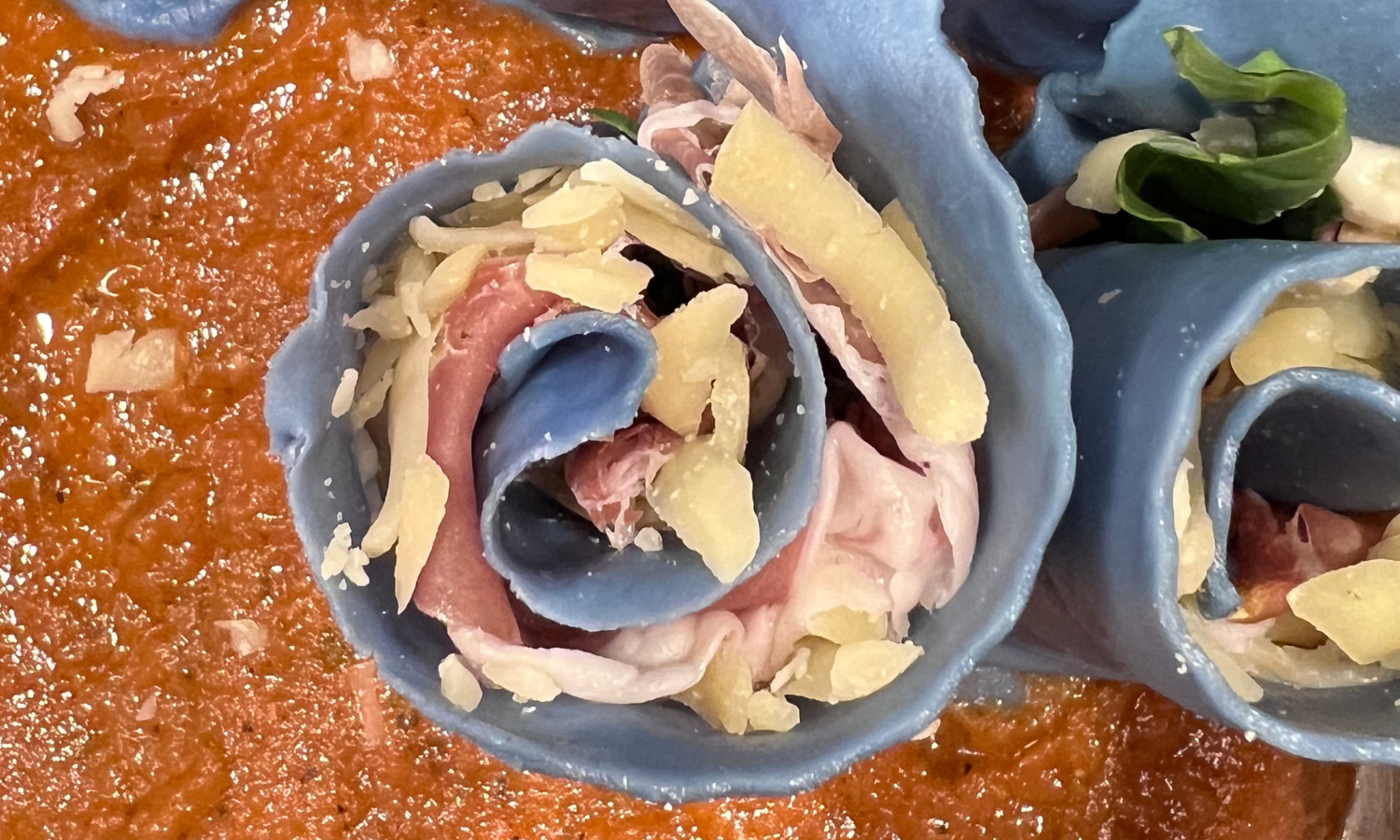 Close-up of decorative blue pottery cups filled with colorful, thinly sliced cured meats, shredded cheese, and garnished with herbs and vegetables, set on a table with a red sauce or dip.
