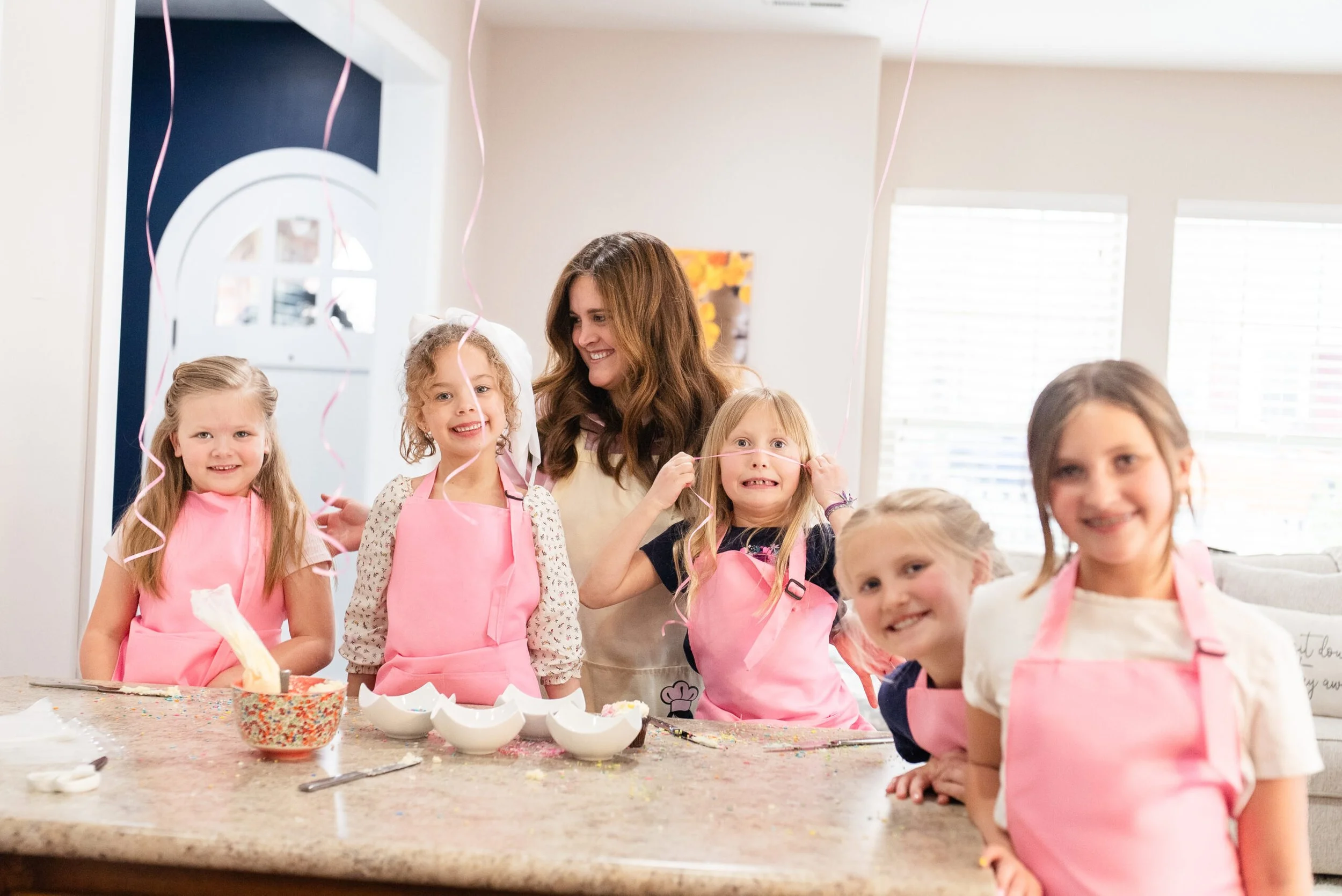 A group of six smiling children and a woman in a kitchen celebrating a birthday with pink aprons, balloons, and cake decorations.
