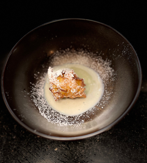 A black bowl containing a piece of fried food coated with powdered sugar on a creamy sauce with additional powdered sugar sprinkled around.