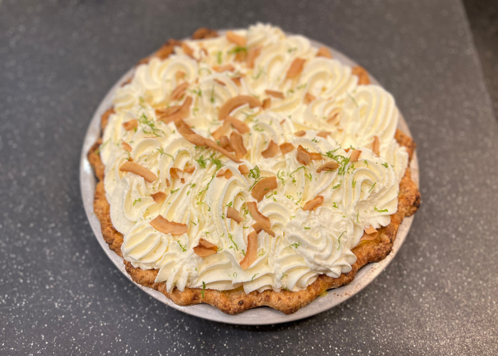 A round nut tart topped with piped whipped cream, toasted coconut flakes, and lime zest on a dark speckled surface.