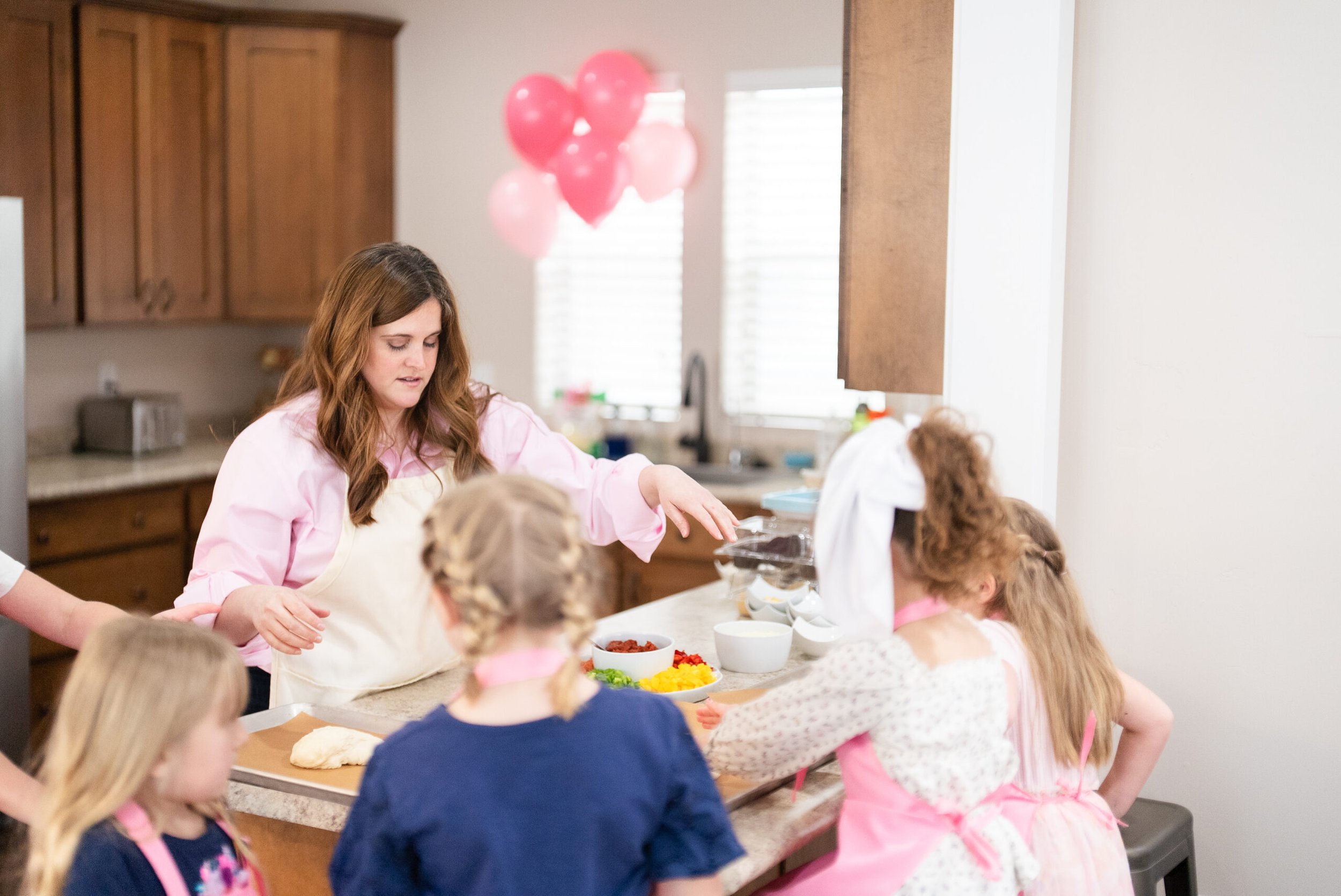 A woman in a pink shirt and apron demonstrates baking to a group of children in a kitchen, with pink balloons in the background.