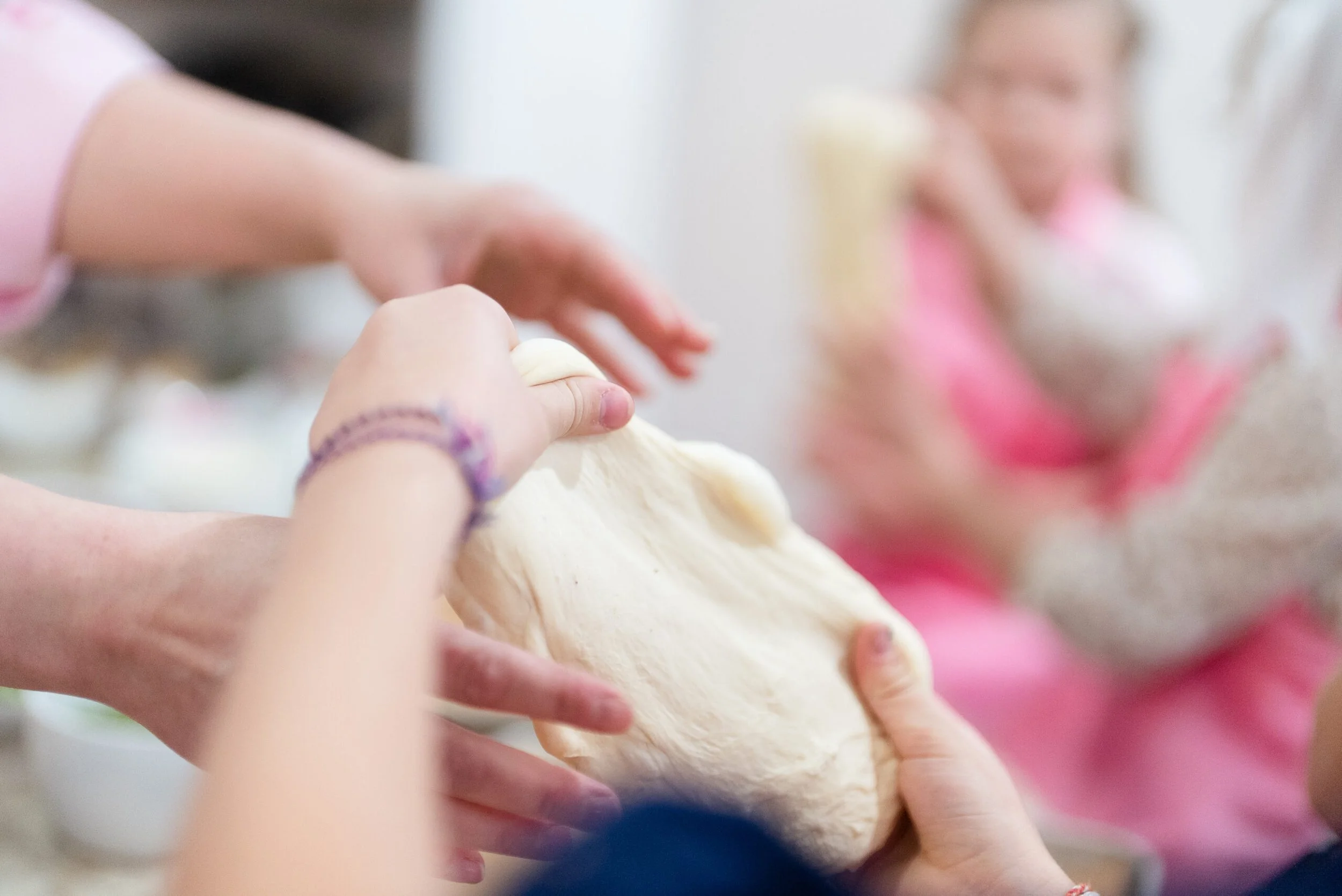Children kneading dough together in a kitchen, with a woman smiling in the background.