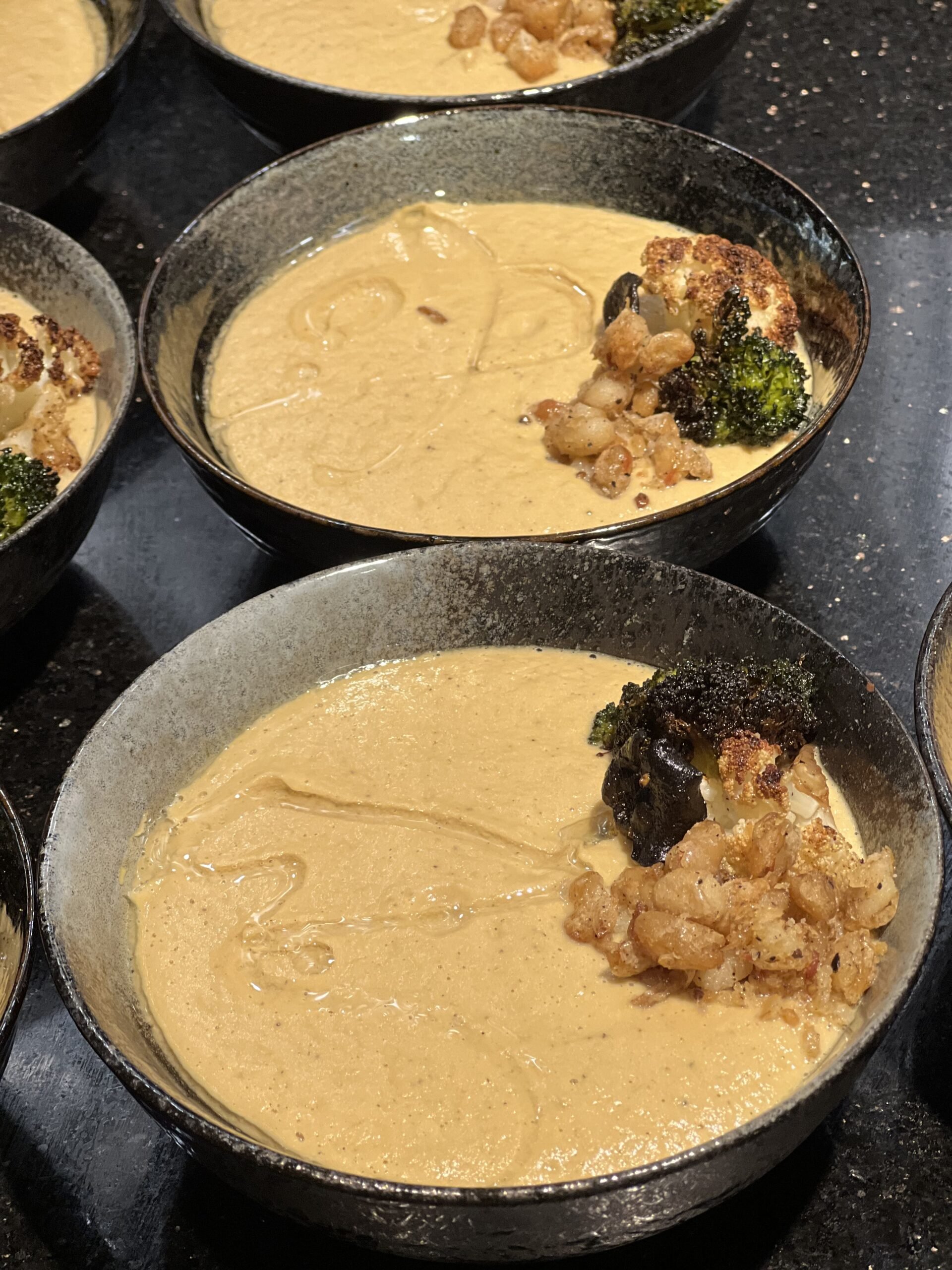 Bowls of creamy soup garnished with roasted broccoli, crumbled bacon, and what appears to be cauliflower.