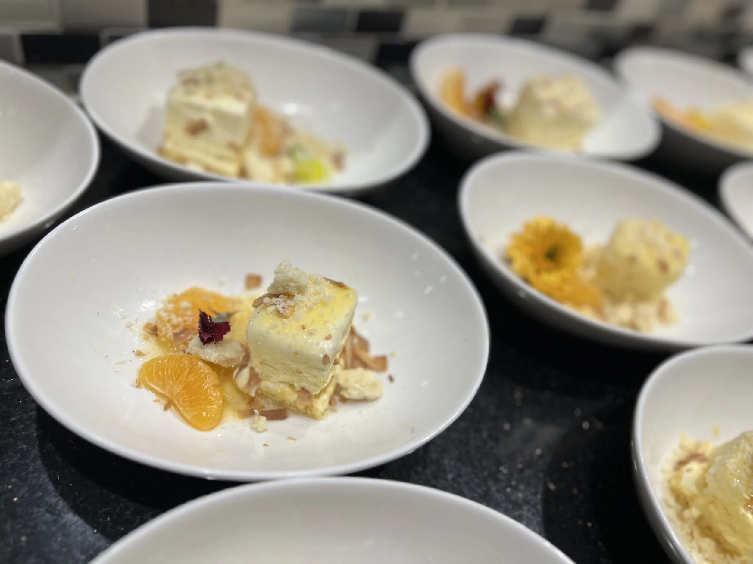 Several white bowls containing a dessert with slices of orange, a small piece of cake, and garnished with edible flowers and crumbled toppings.
