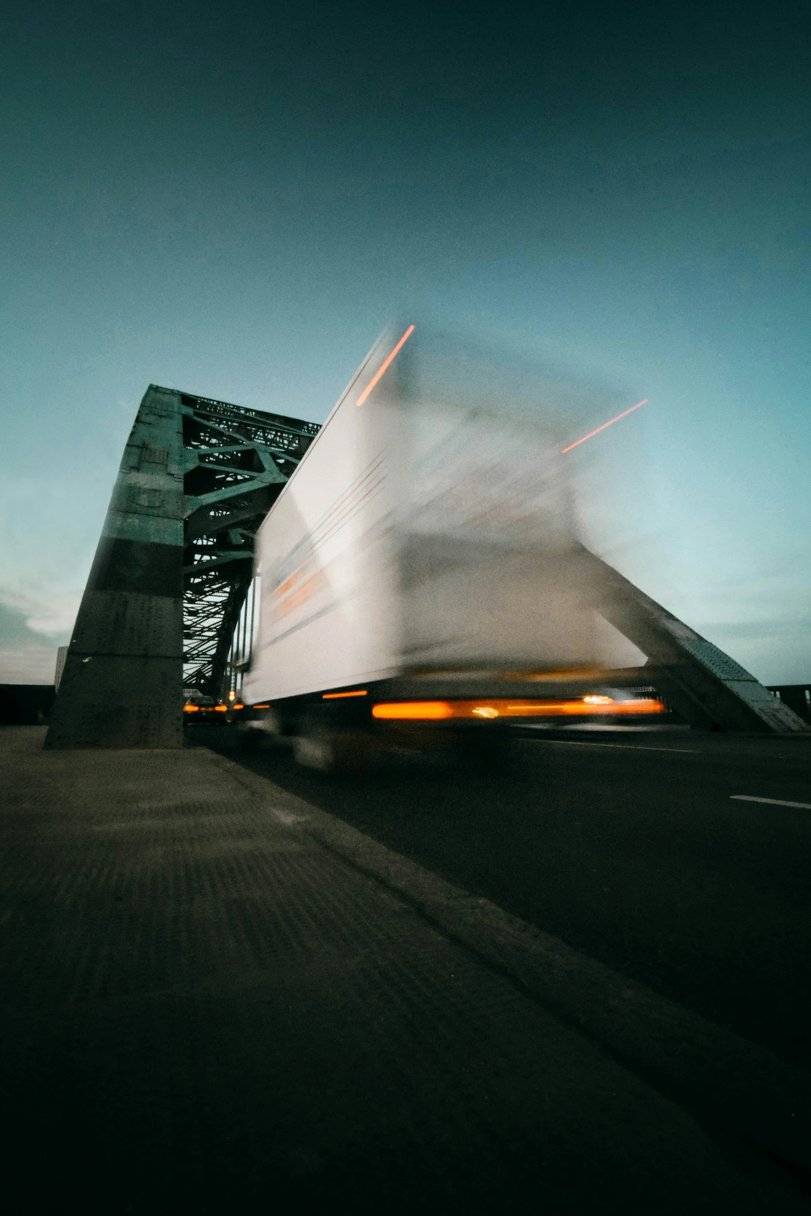 Blurred truck speeding over a bridge at dusk, with a structure supporting the bridge visible on the left.