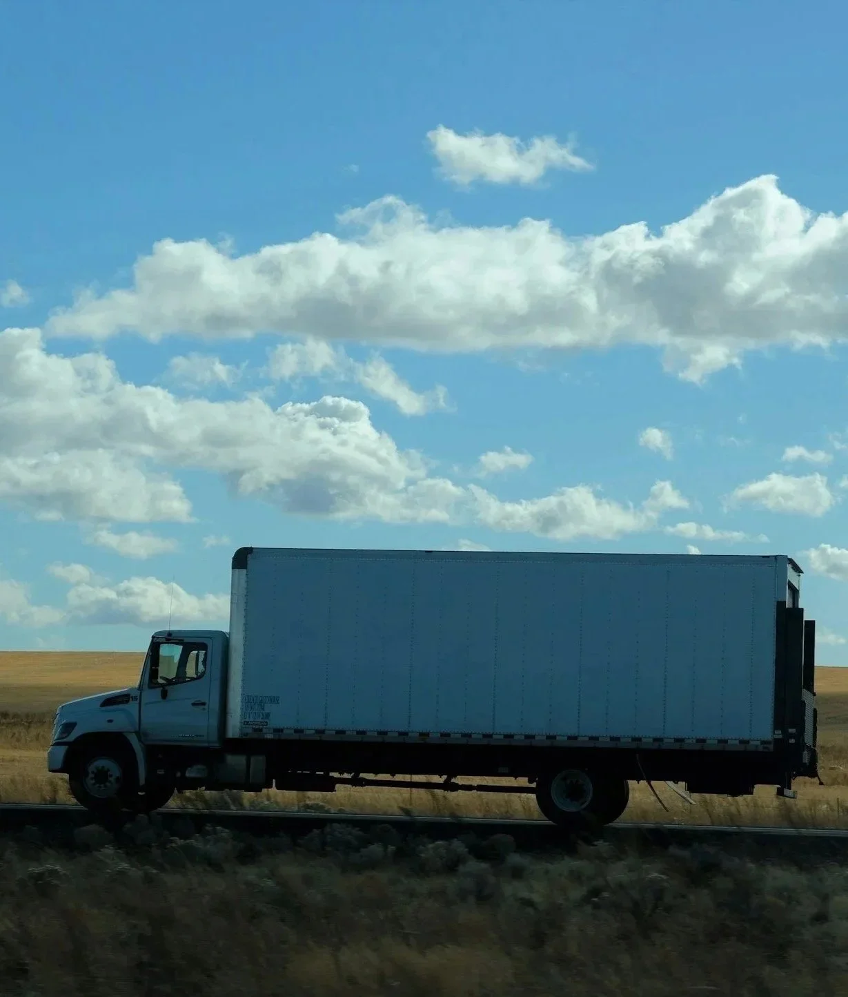 A large white delivery truck driving on a railroad track through a rural landscape with grassy fields and a partly cloudy sky.