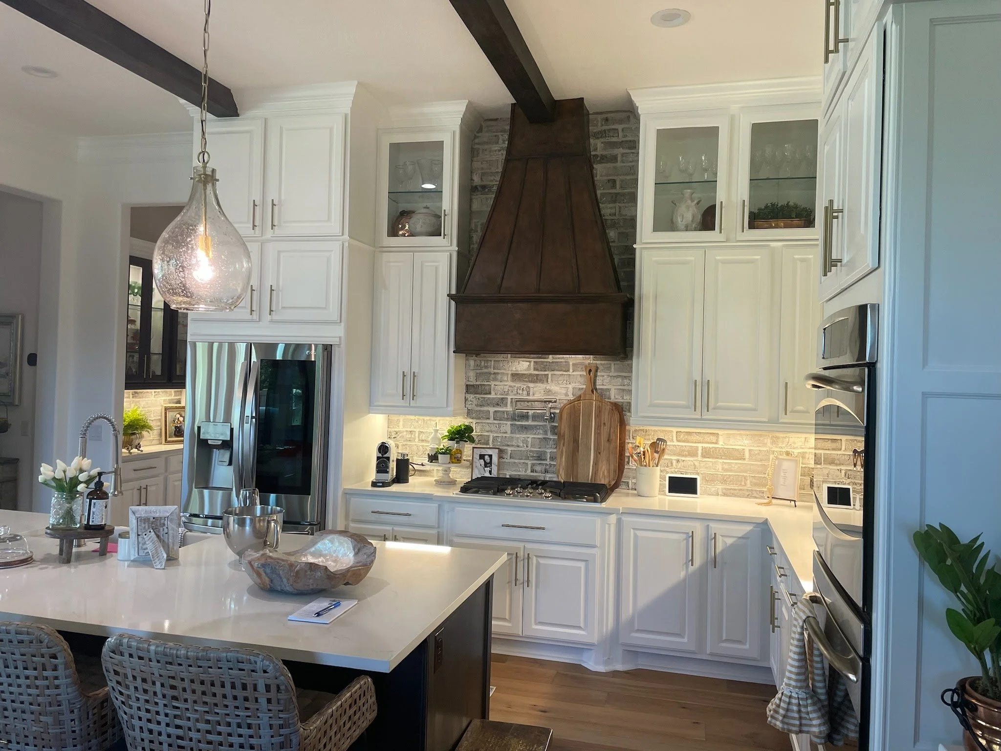 Modern kitchen with white cabinets, a kitchen island with a white countertop, stainless steel appliances, a wooden range hood, and decorative items including plants and jars.