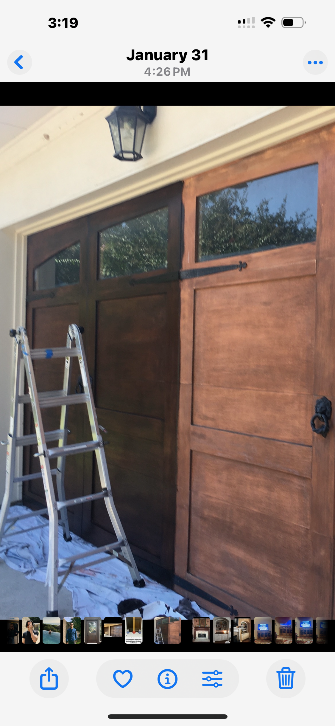 A photo of a wooden garage door under renovation, with a ladder and painting supplies on a white tarp below, and a black outdoor lantern mounted above.