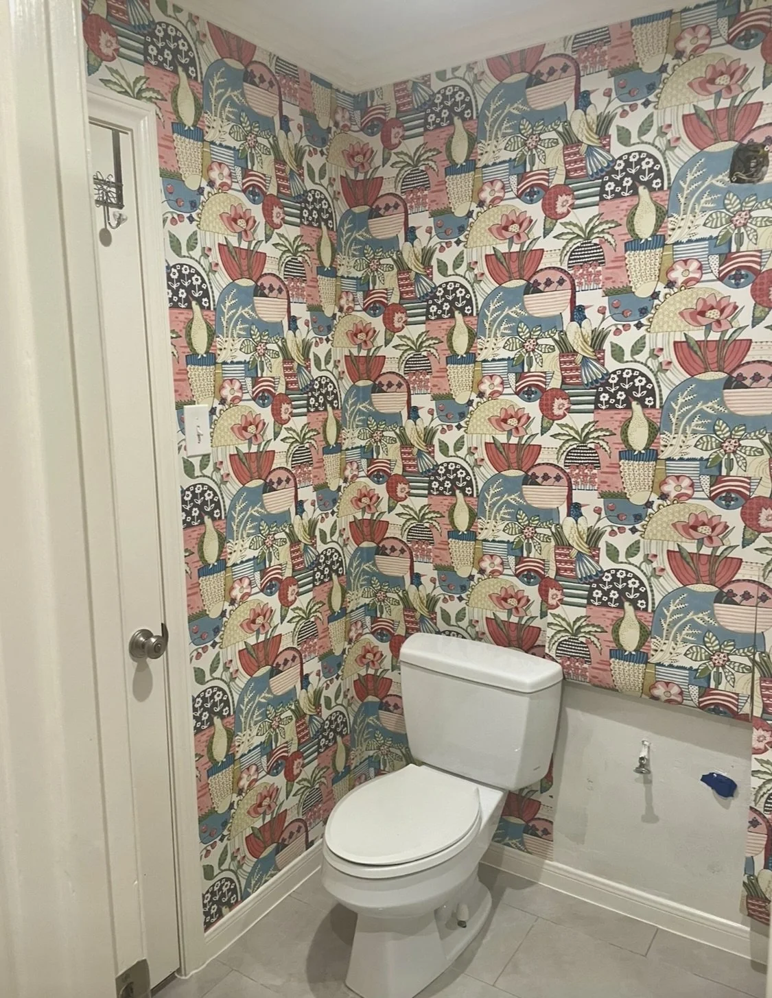 A bathroom with vibrant wallpaper featuring large tropical flowers, leaves, and abstract shapes in pink, blue, green, and black. A white toilet is positioned against the wall, and there are wall fixtures and outlets nearby.