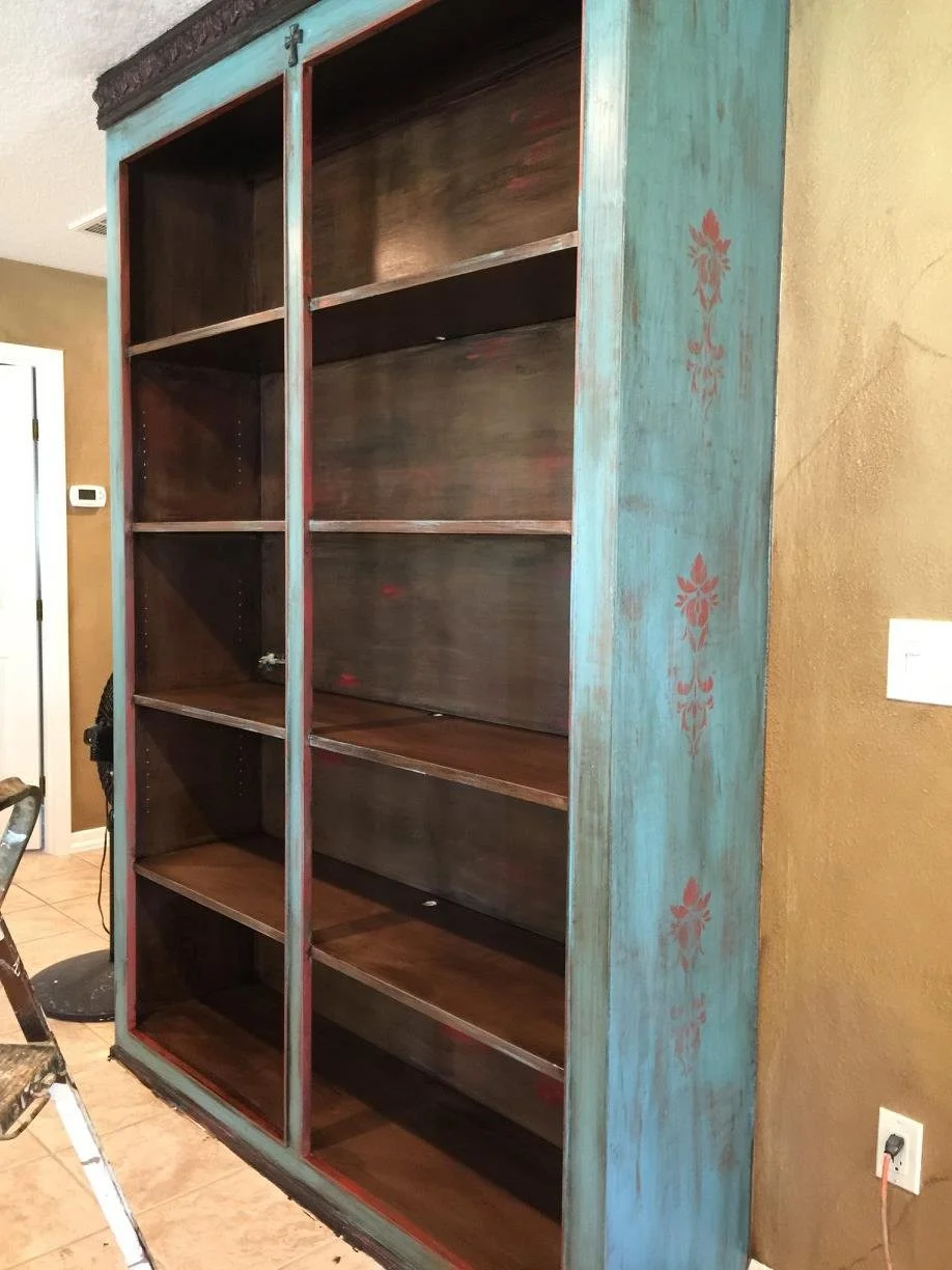 A tall wooden bookshelf with a distressed blue frame with red flower patterns on the side, standing against a beige wall in a room.