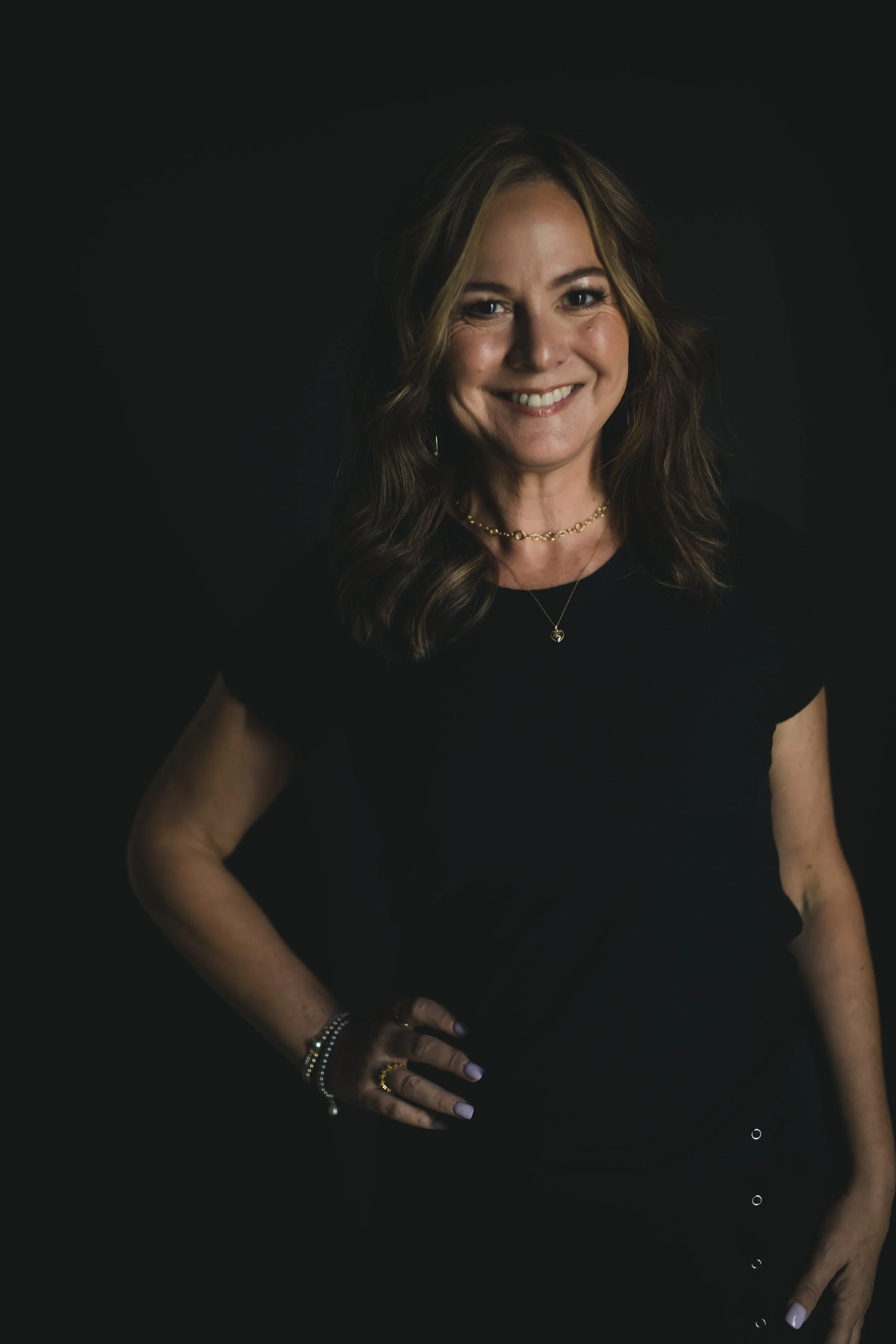 A woman with long, wavy brown hair smiling, wearing a black top, gold jewelry, and light purple nail polish, standing against a dark background.