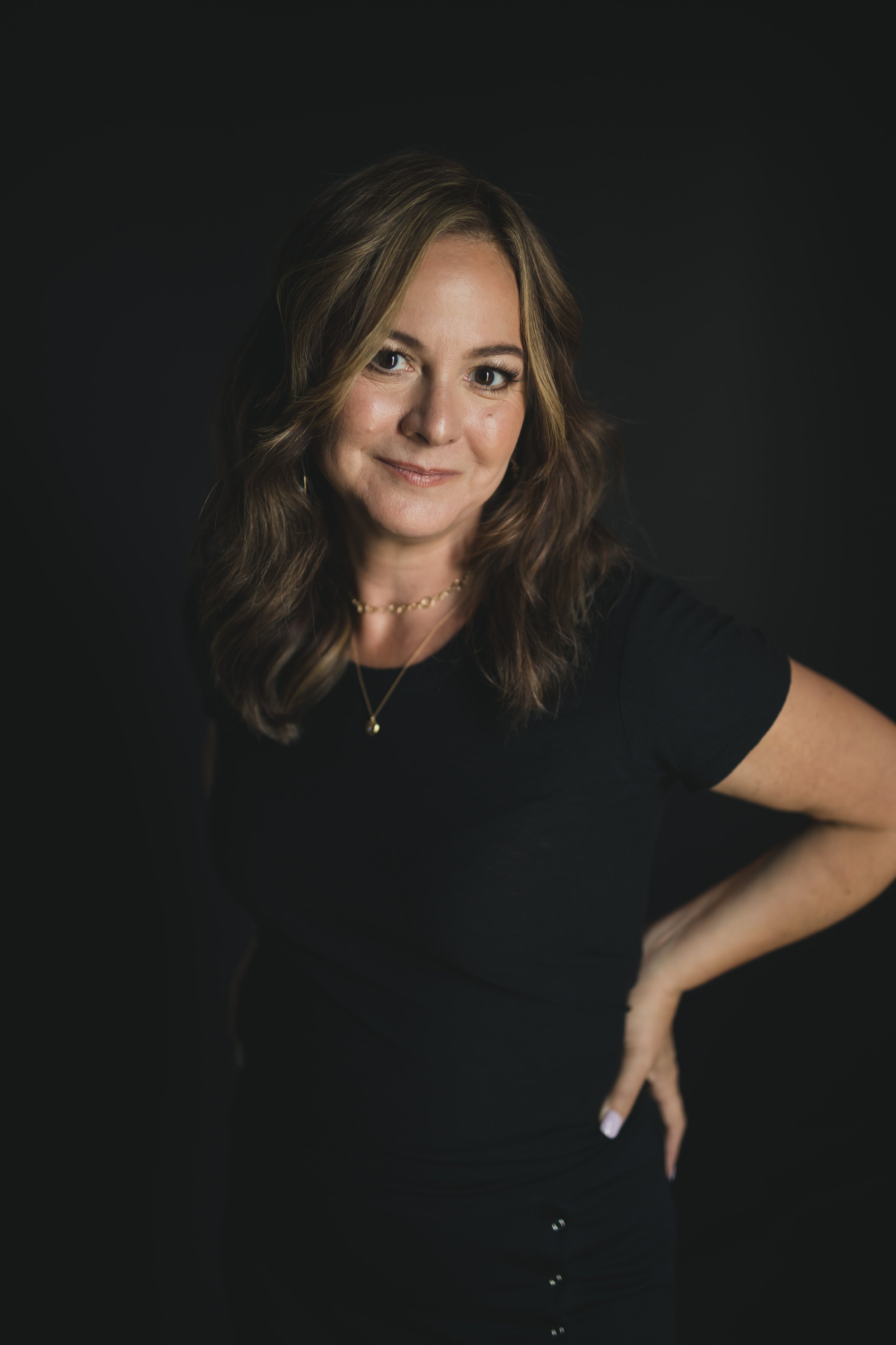 Michelle Sheppard. A woman with wavy brown hair, wearing a black t-shirt and gold jewelry, standing against a black background, looking at the camera with a slight smile.