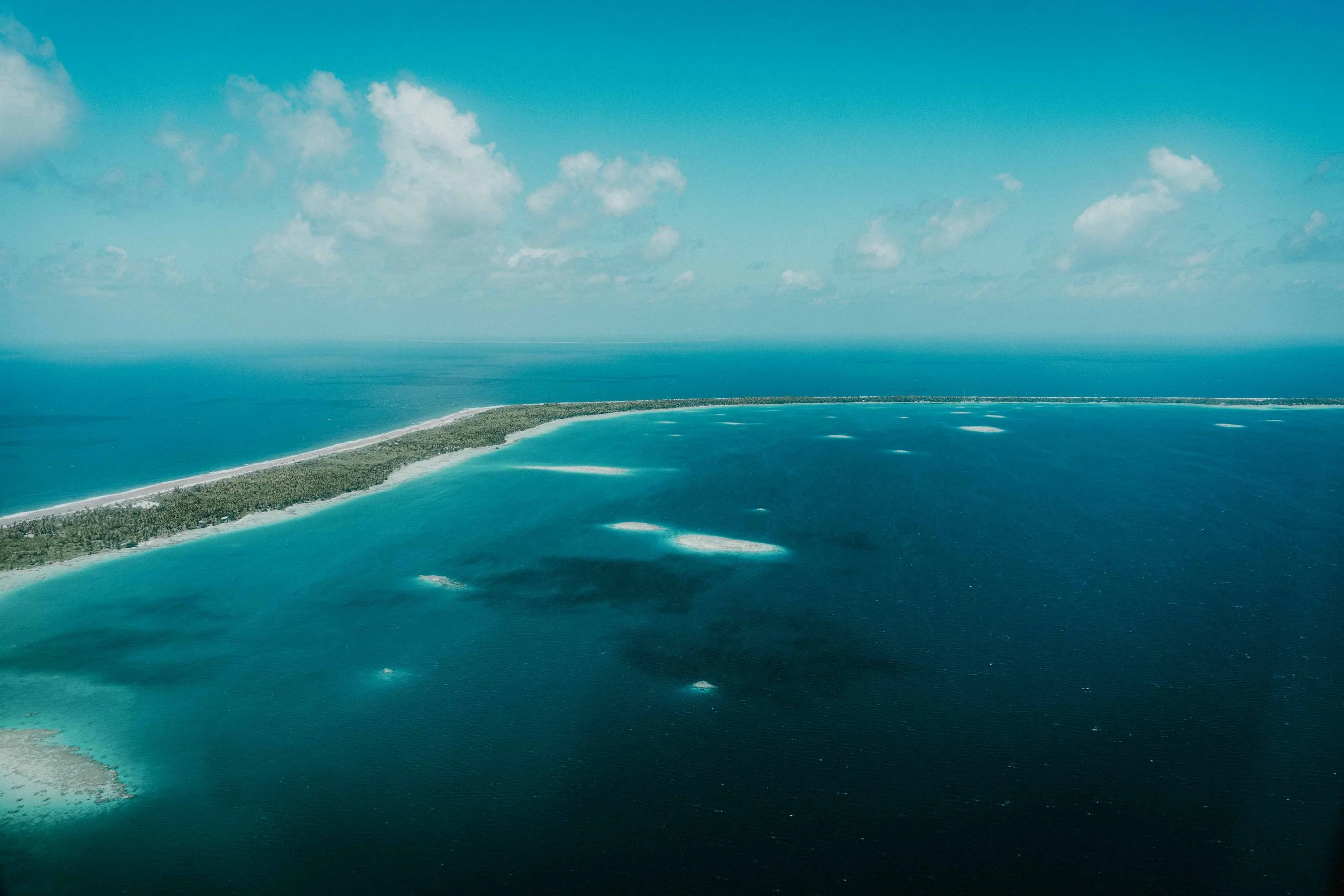 Vue aérienne d'une barrière de corail au bord de l'océan, avec des lagons turquoise et une étendue d'eau profonde, sous un ciel bleu avec quelques nuages.
