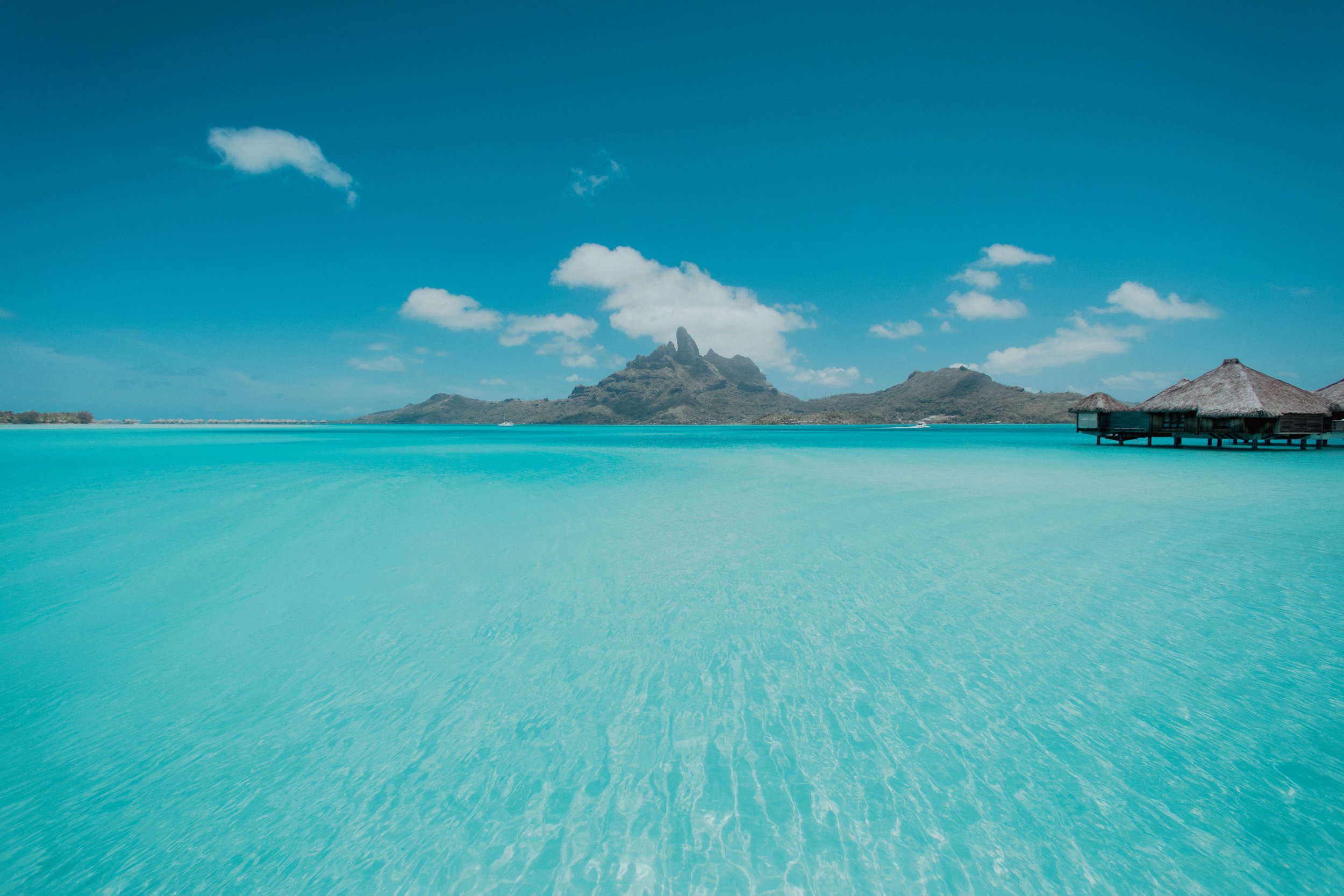 Une plage de sable blanc avec une eau turquoise claire, des cabanes sur pilotis à droite et une montagne avec un sommet rocheux en arrière-plan sous un ciel bleu avec quelques nuages.