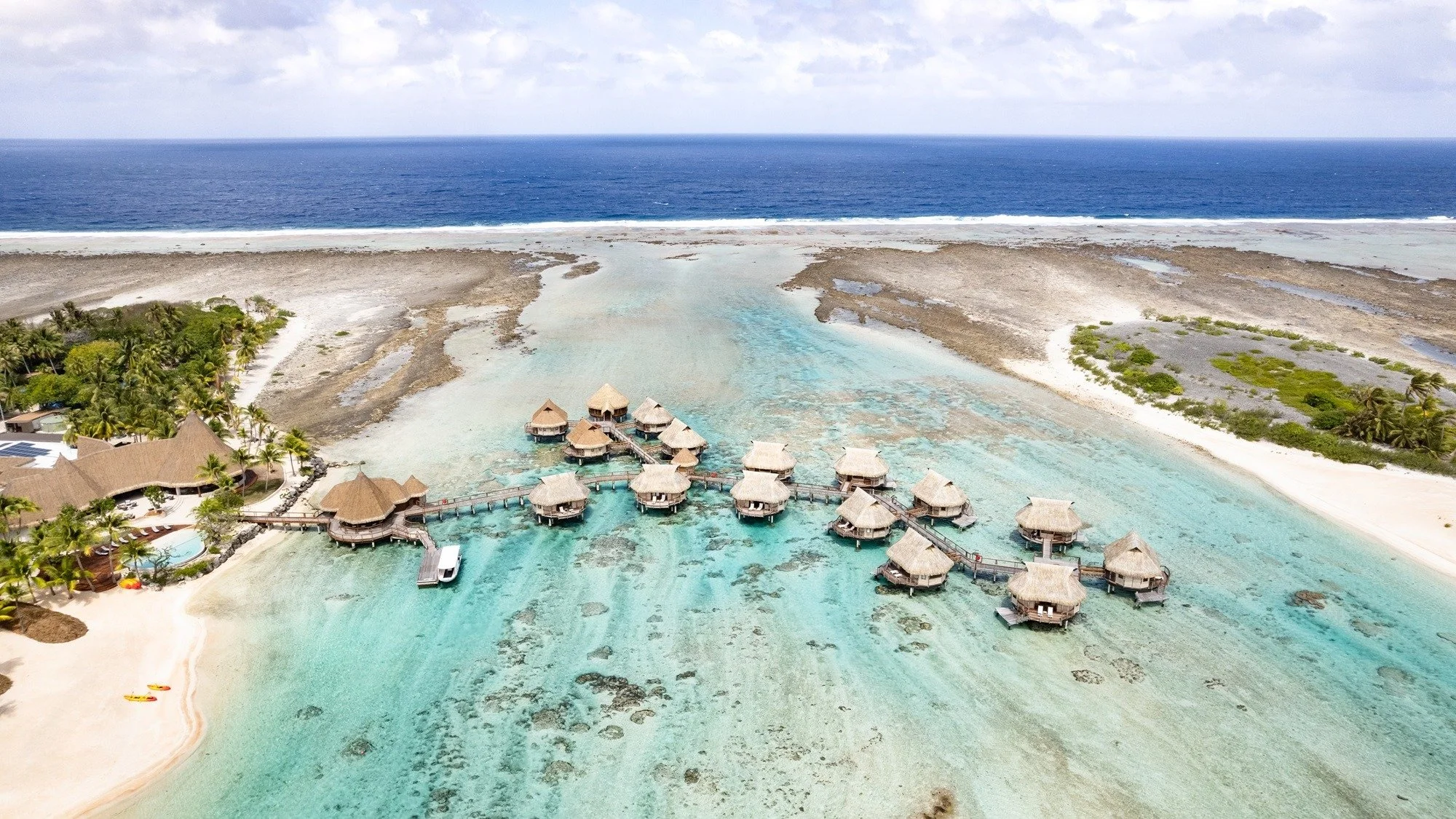 Vue aérienne d'un complexe hôtelier avec des bungalows en surplomb sur l'eau, entouré d'une plage de sable blanc et de végétation tropicale, avec l'océan au loin.