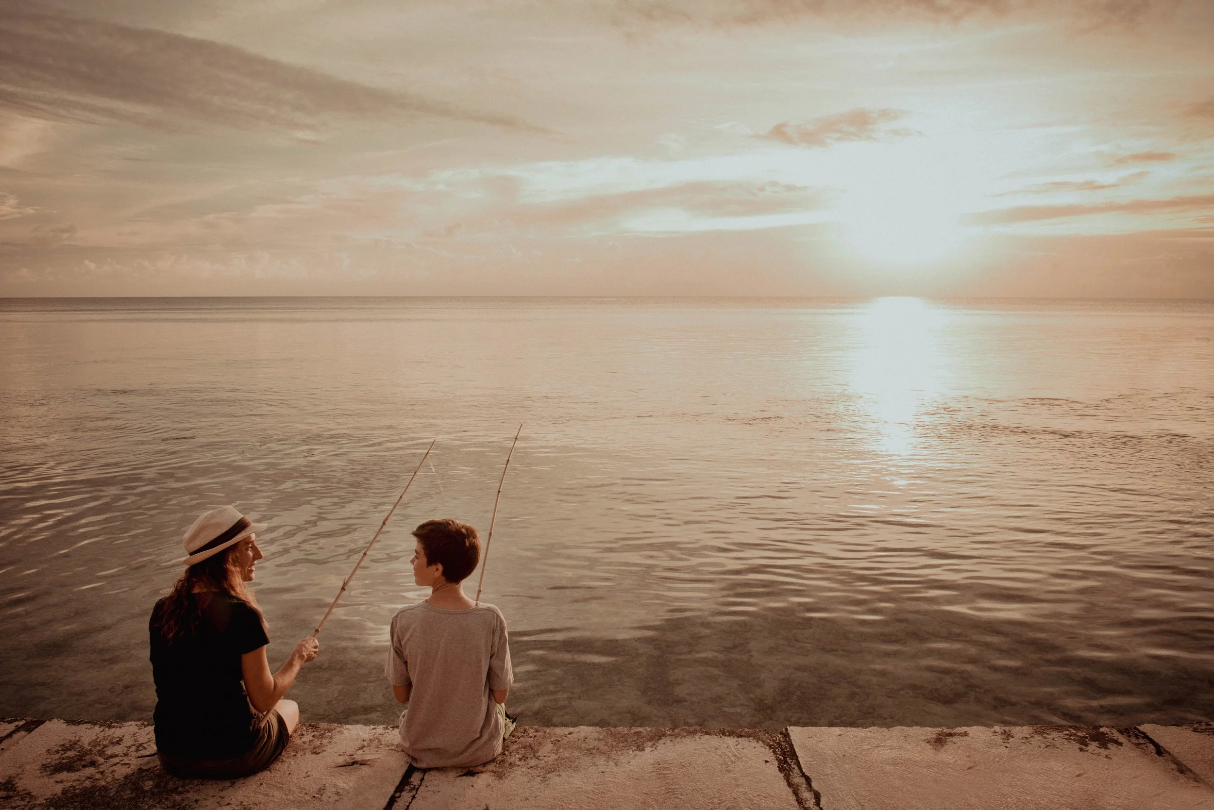 Une femme et un enfant assis sur un quai, en train de pêcher au coucher du soleil sur une mer calme.