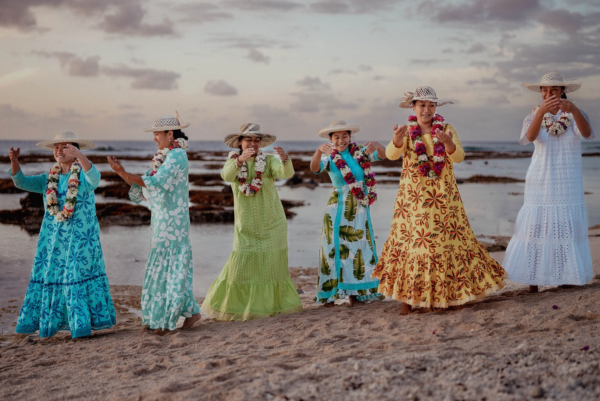 Groupe de femmes en robes colorées, portant des chapeaux de paille et des colliers de fleurs, qui dansent sur la plage au coucher du soleil.