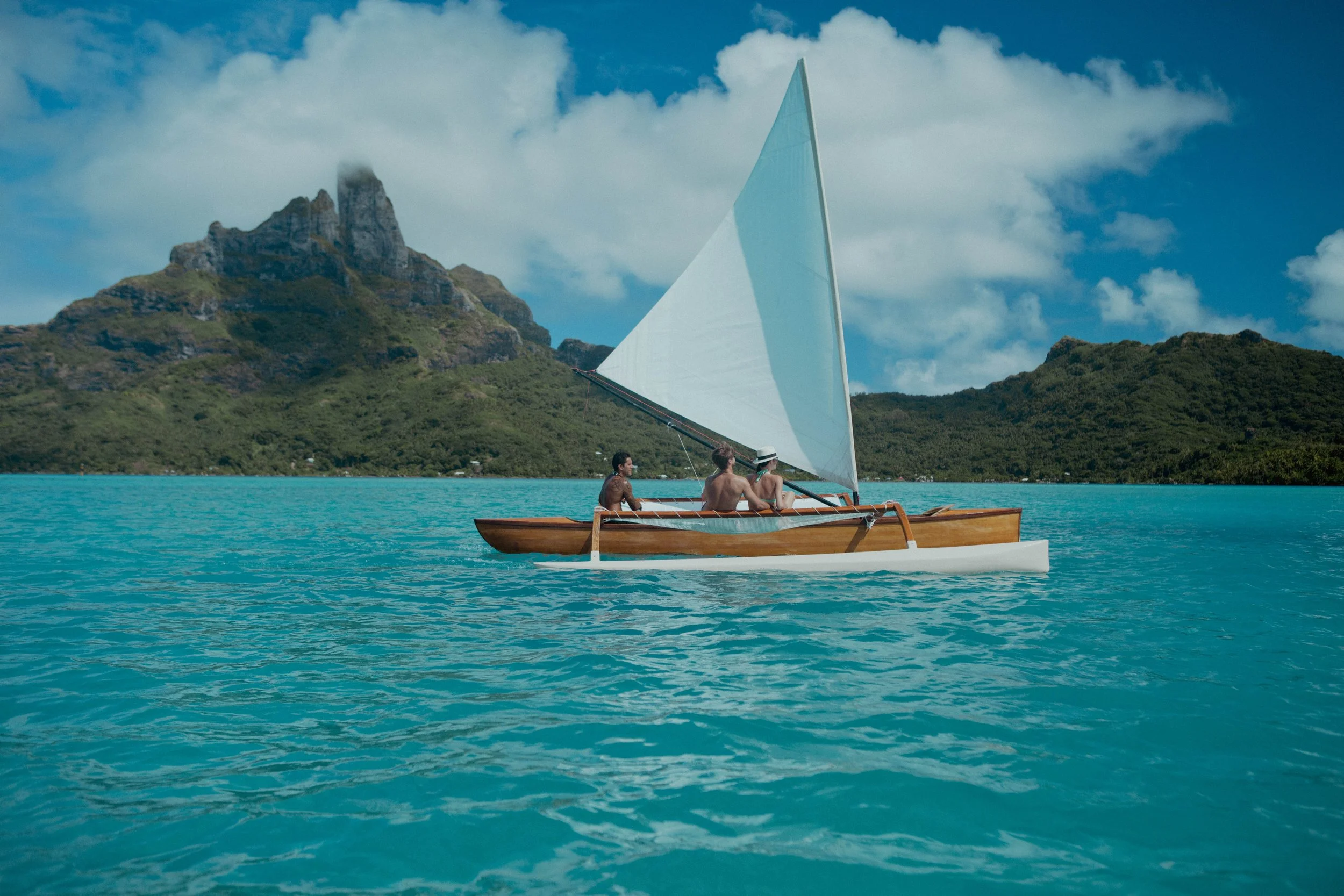 Trois personnes dans un bateau à voile en pleine mer turquoise avec des montagnes verdoyantes en arrière-plan, sous un ciel partiellement nuageux.