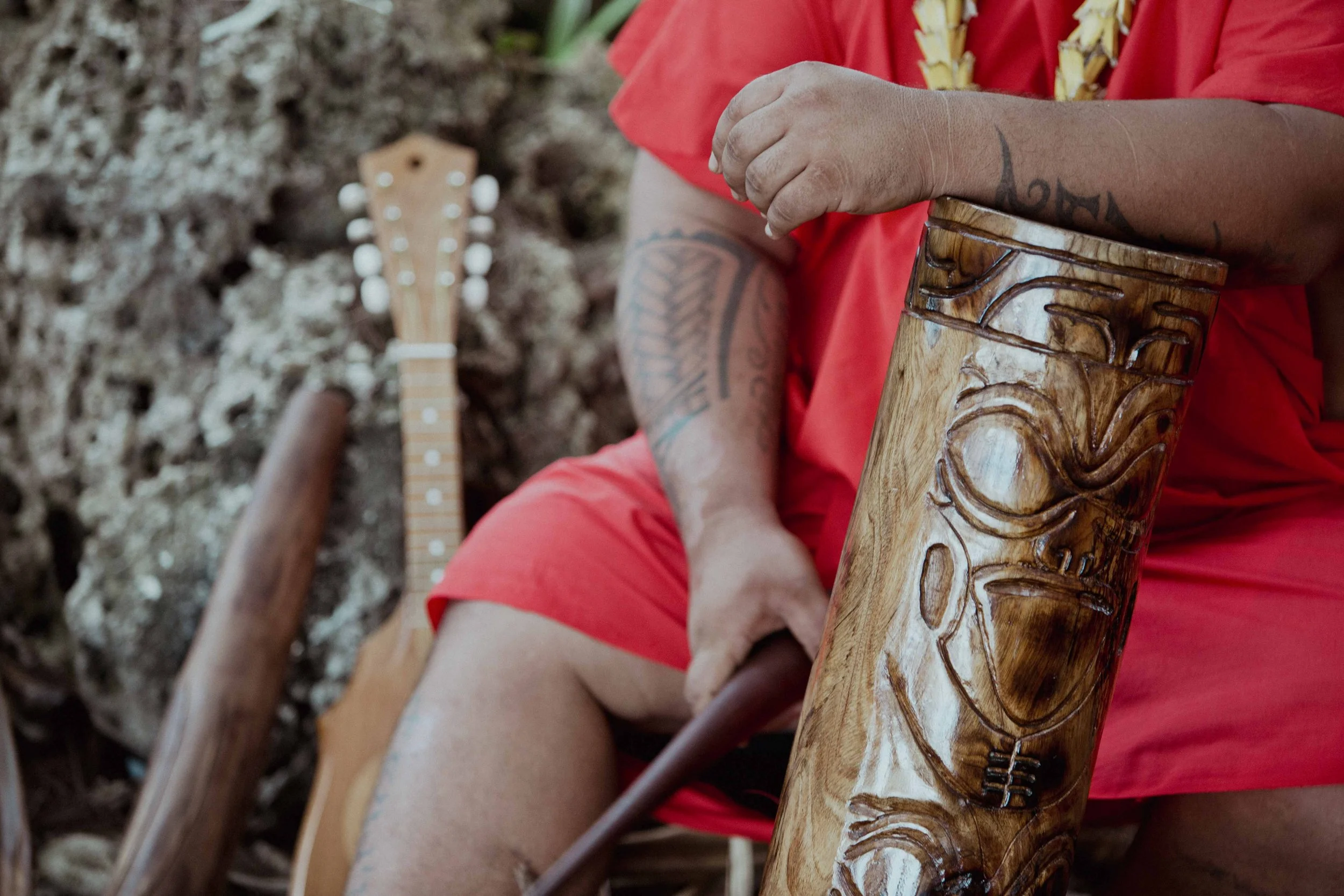 Personne avec tatouages jouant de la musique avec une flûte en bois sculptée et un ukulélé, en plein air, vêtue d'une robe rouge.