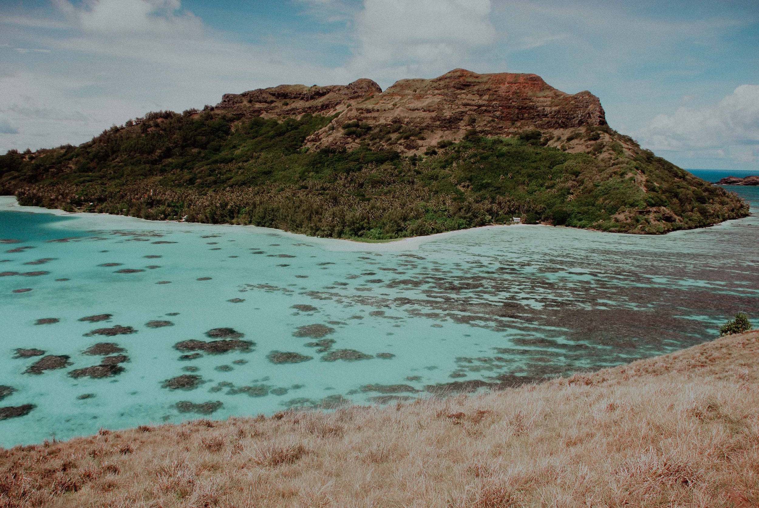 Une île avec une montagne verte entourée d'eau turquoise, avec une plage de sable claire au premier plan.