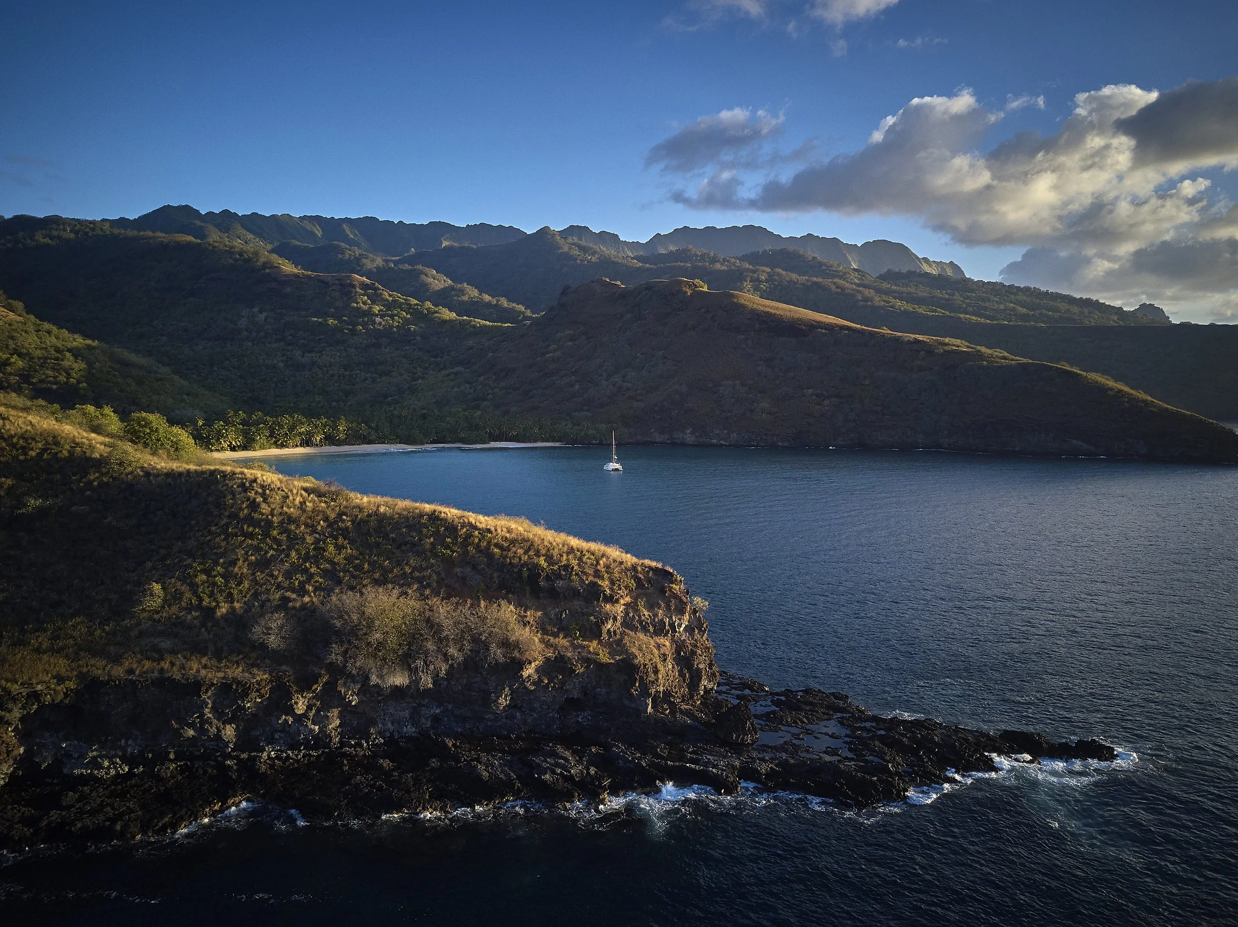 Coucher de soleil sur une baie entourée de montagnes verdoyantes, avec un seul bateau à voile sur l'eau calme, et une côte rocheuse en premier plan.