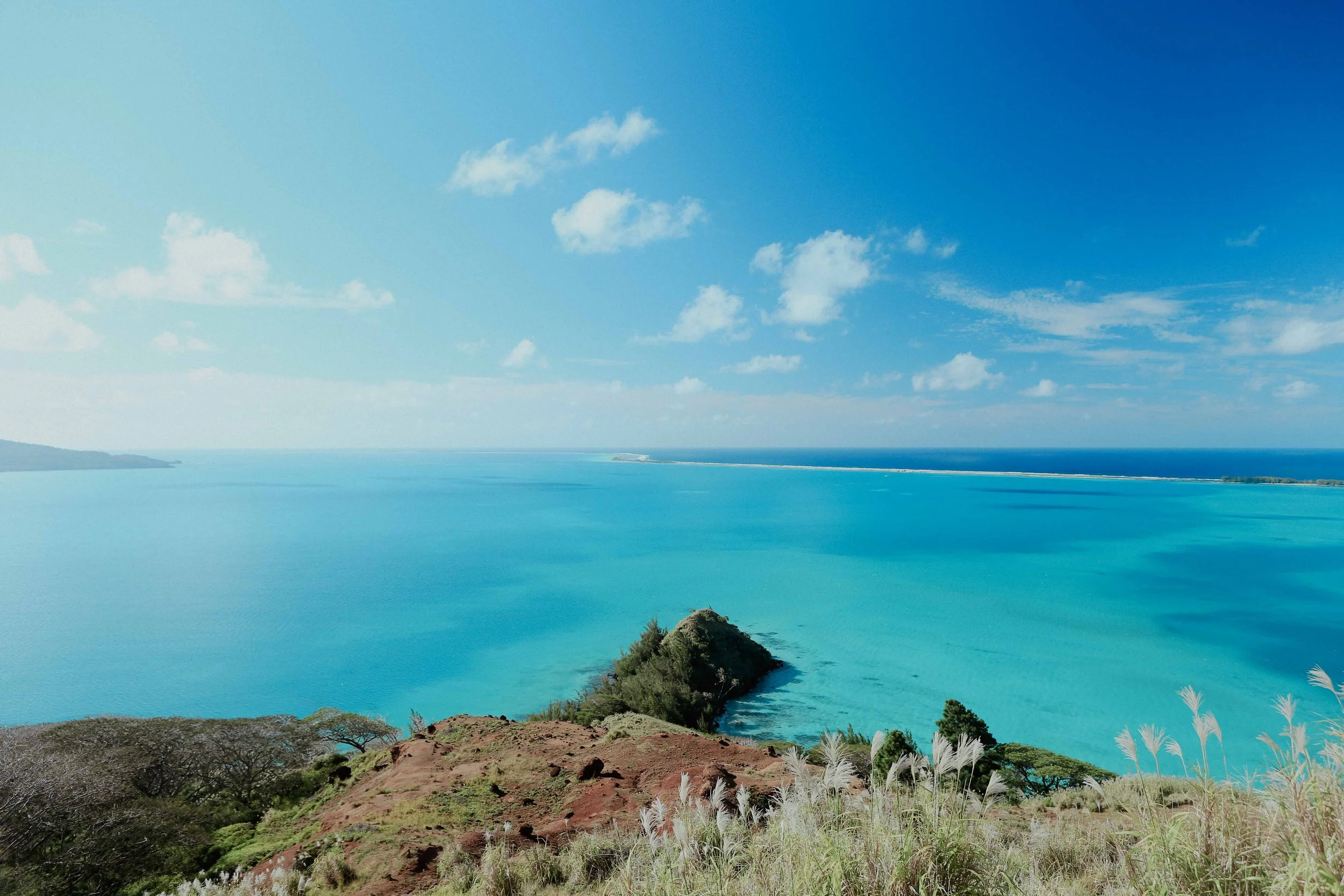 Une vue panoramique d'une mer turquoise avec un rocher au centre, entourée de végétation verte et de collines, sous un ciel bleu clair avec quelques nuages.